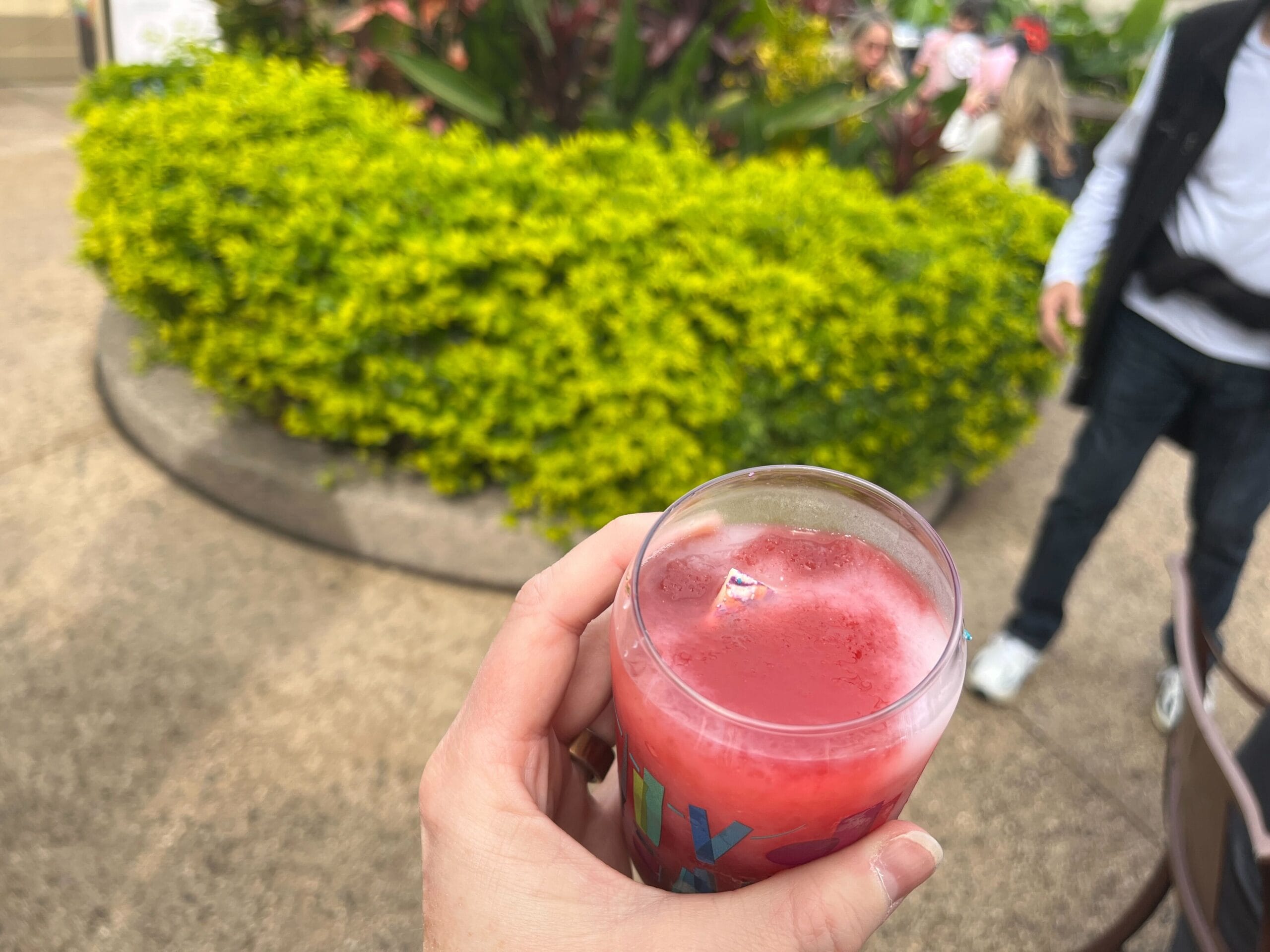 A hand holds a partially consumed frozen red drink in a plastic cup, capturing the essence of the Festival of the Arts. In the background, a vibrant green bush and a person walking add to the lively scene.