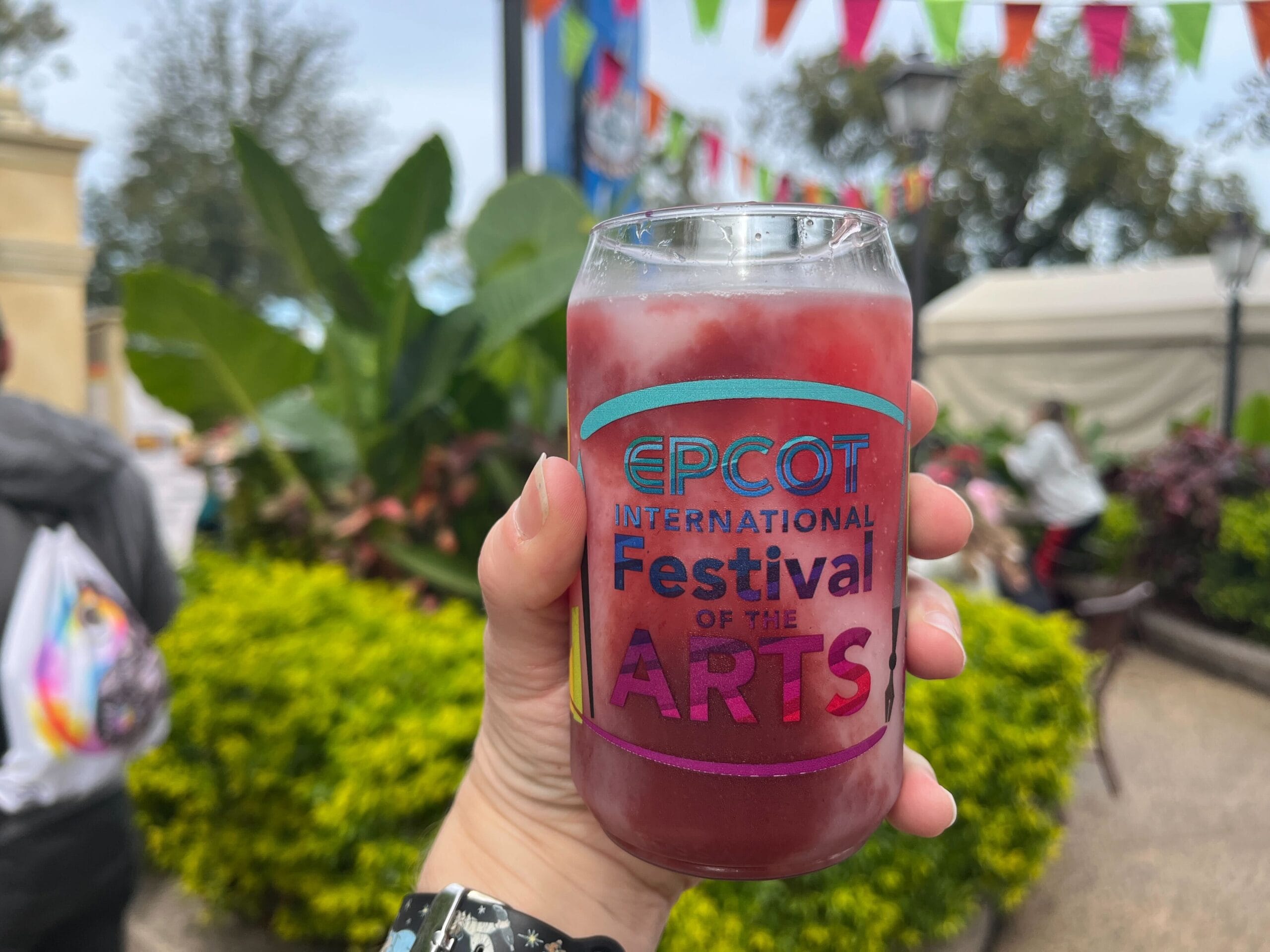 A hand gracefully holds a glass with a vibrant drink, marked "EPCOT International Festival of the Arts." The scene is adorned with festive decorations and lush greenery, capturing the creative spirit of the Festival of the Arts.