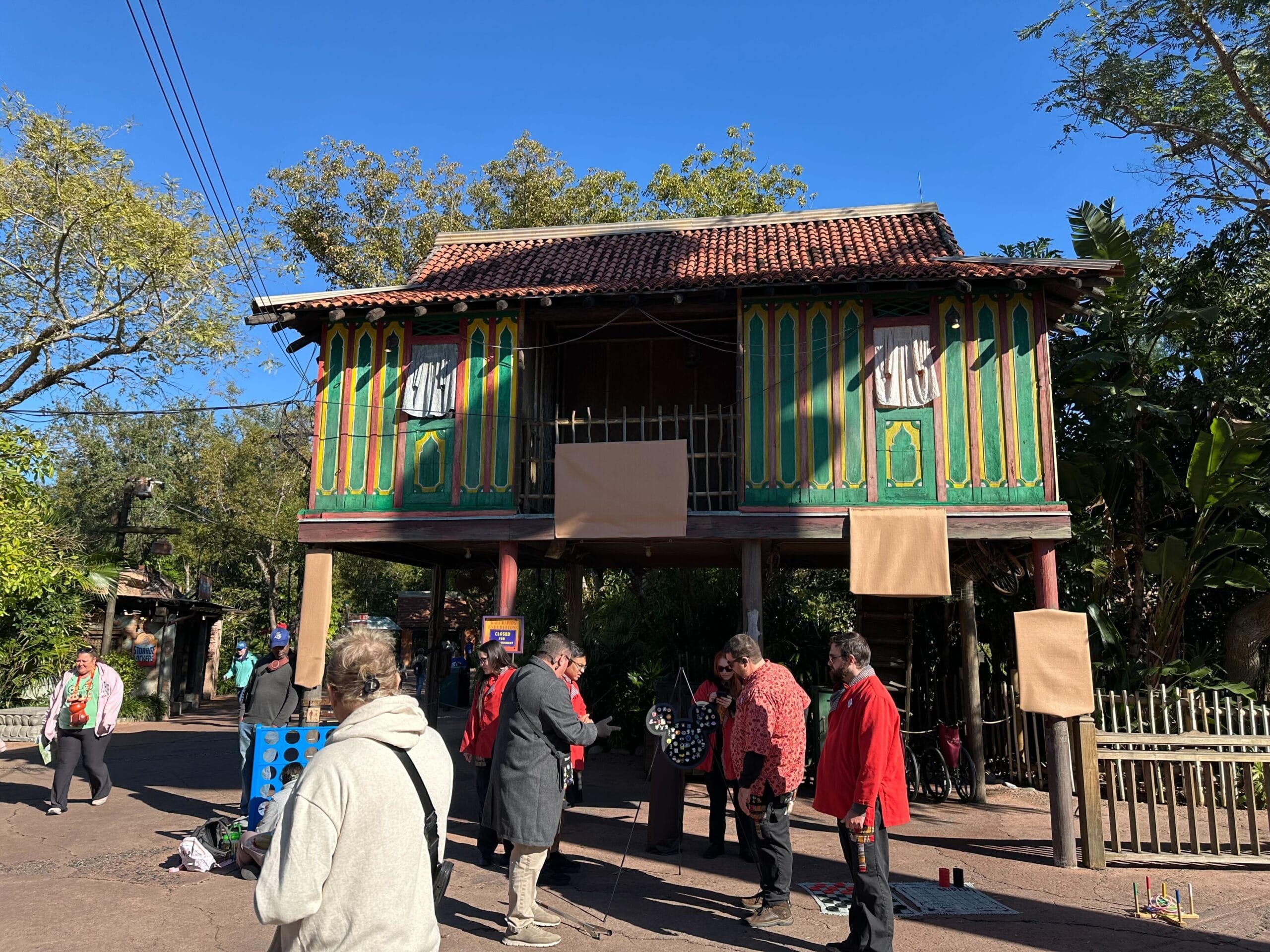 People gathered near a colorful, elevated wooden house with a tiled roof. Trees surround the area, and a clear blue sky is visible.