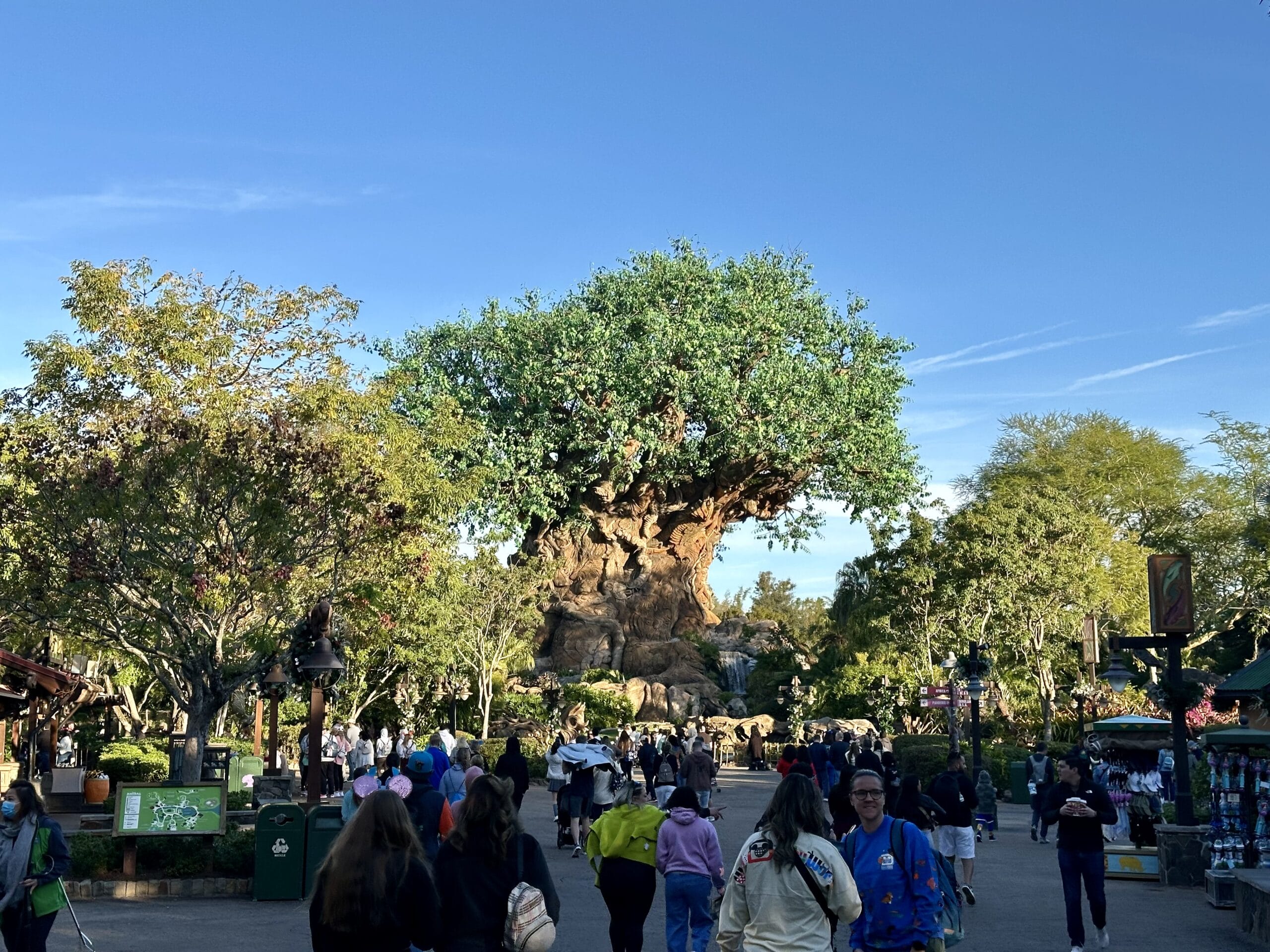 People walking towards a large artificial tree with detailed carvings, surrounded by greenery and clear blue sky.