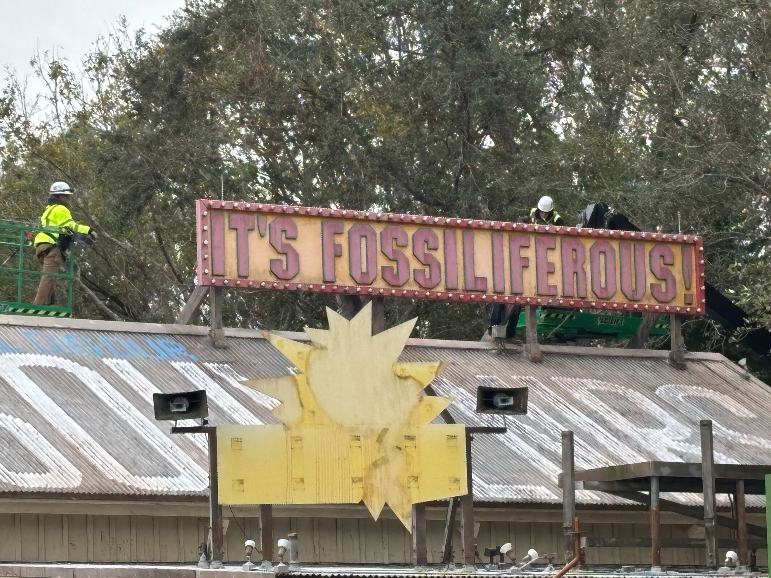 Workers on a roof install or repair a sign that reads "IT'S FOSSILIFEROUS!" amidst trees in the background, capturing the spirit of Dino-Rama.