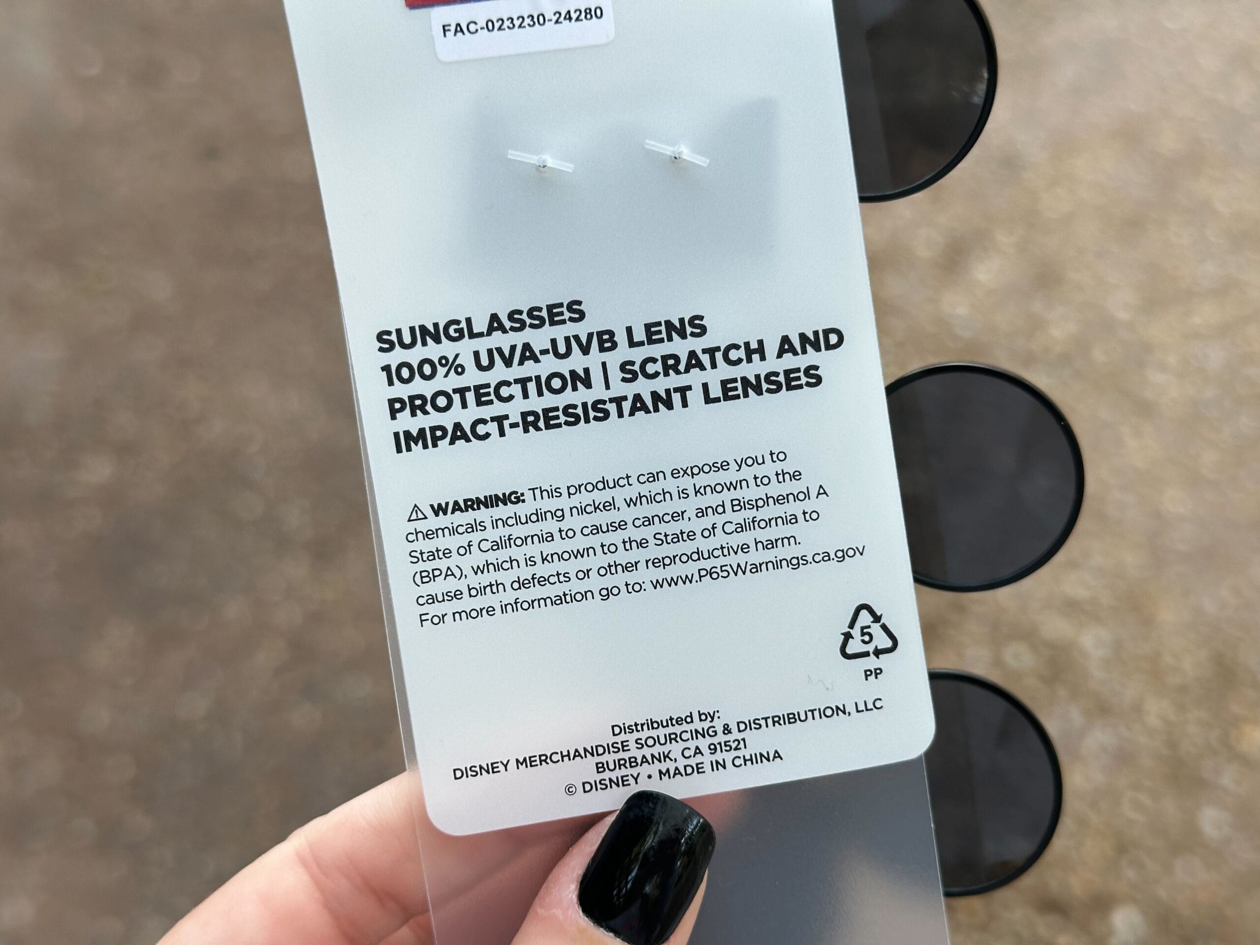 A person holds a set of Mickey Sunglasses with a label describing 100% UVA-UVB lens protection, and scratch and impact resistance. A warning about chemical exposure is included, making them perfect for sunny strolls at Tokyo Disney Resort.