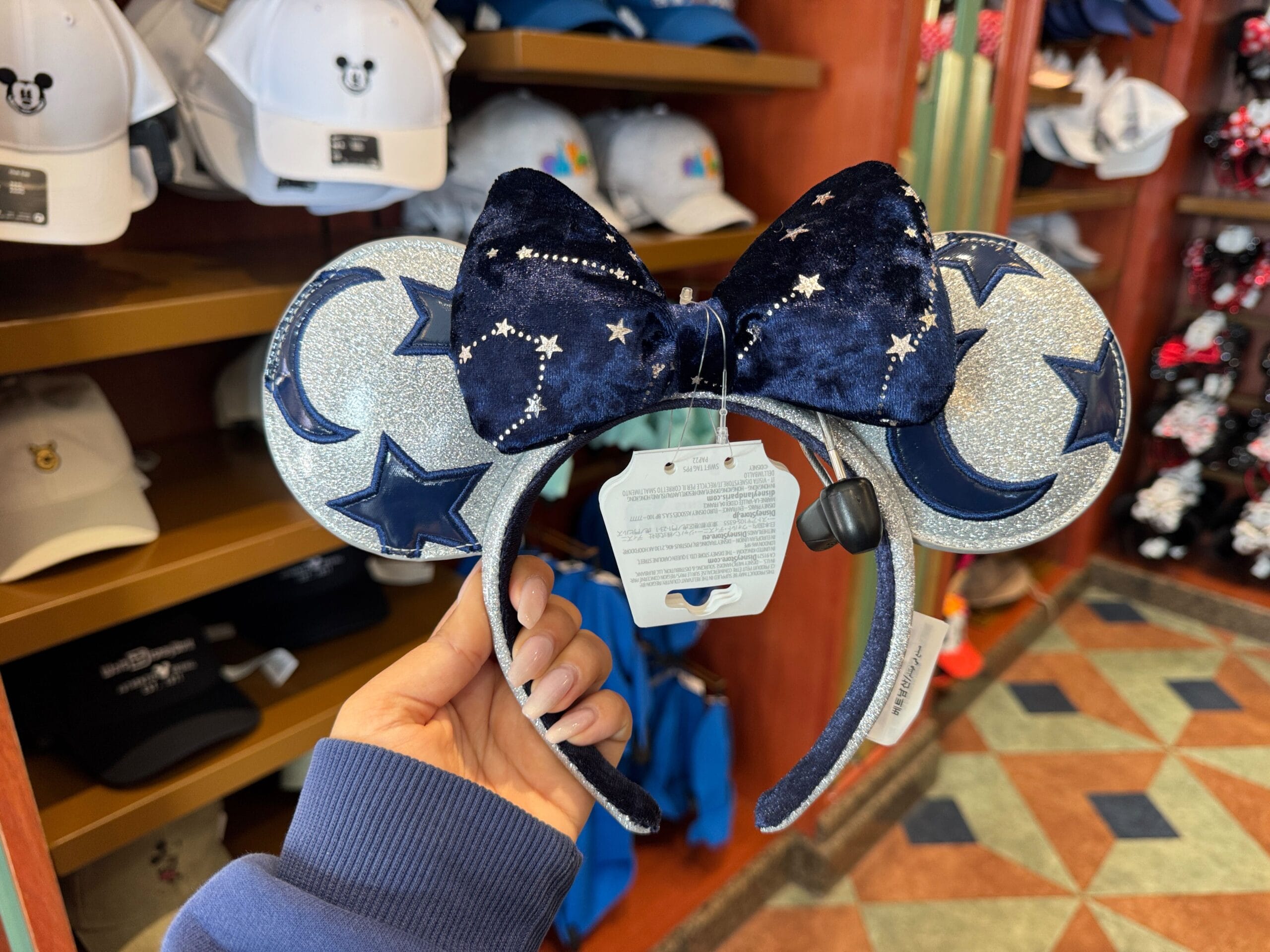 A hand holds a Mickey Mouse-themed headband featuring star-patterned ears and a starry bow, reminiscent of a constellation, displayed in a store with several hats in the background. Perfect for your next Walt Disney World adventure.