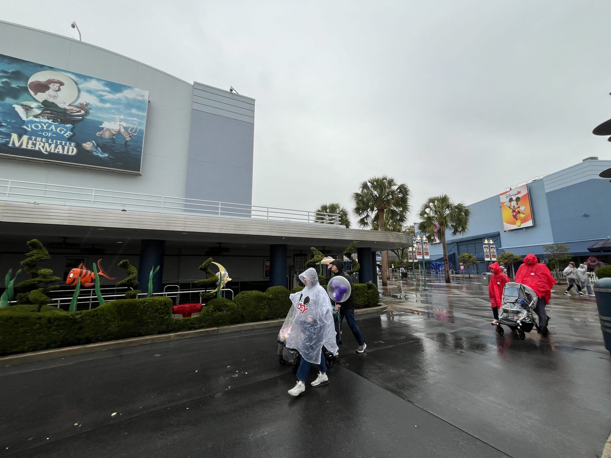 People in raincoats walk on a wet pavement outside a building with the "Voyage of The Little Mermaid" marquee on a cloudy day.