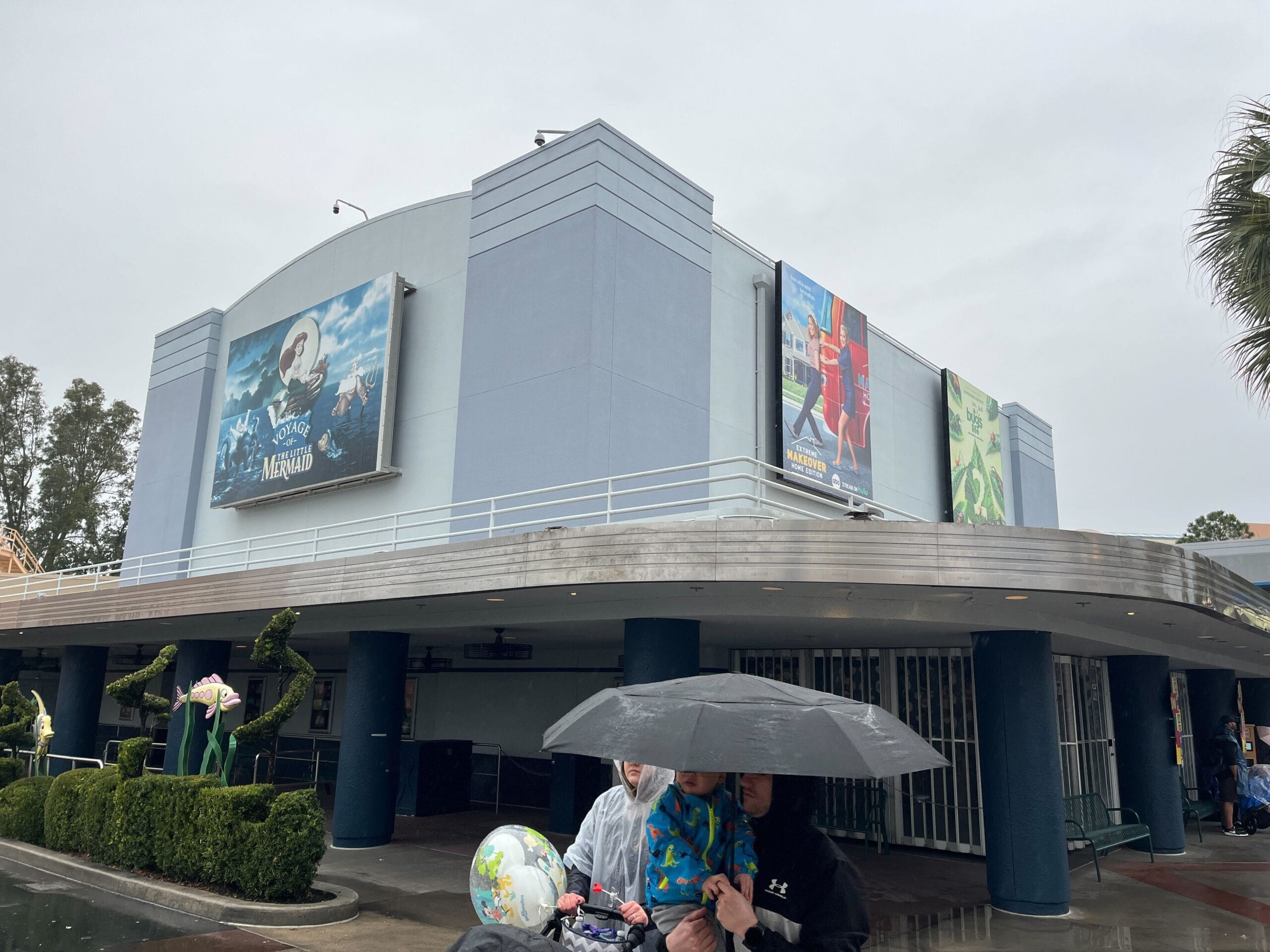 People with an umbrella walk past a building adorned with movie posters, reminiscent of the sights at Disney's Hollywood Studios, on a rainy day.