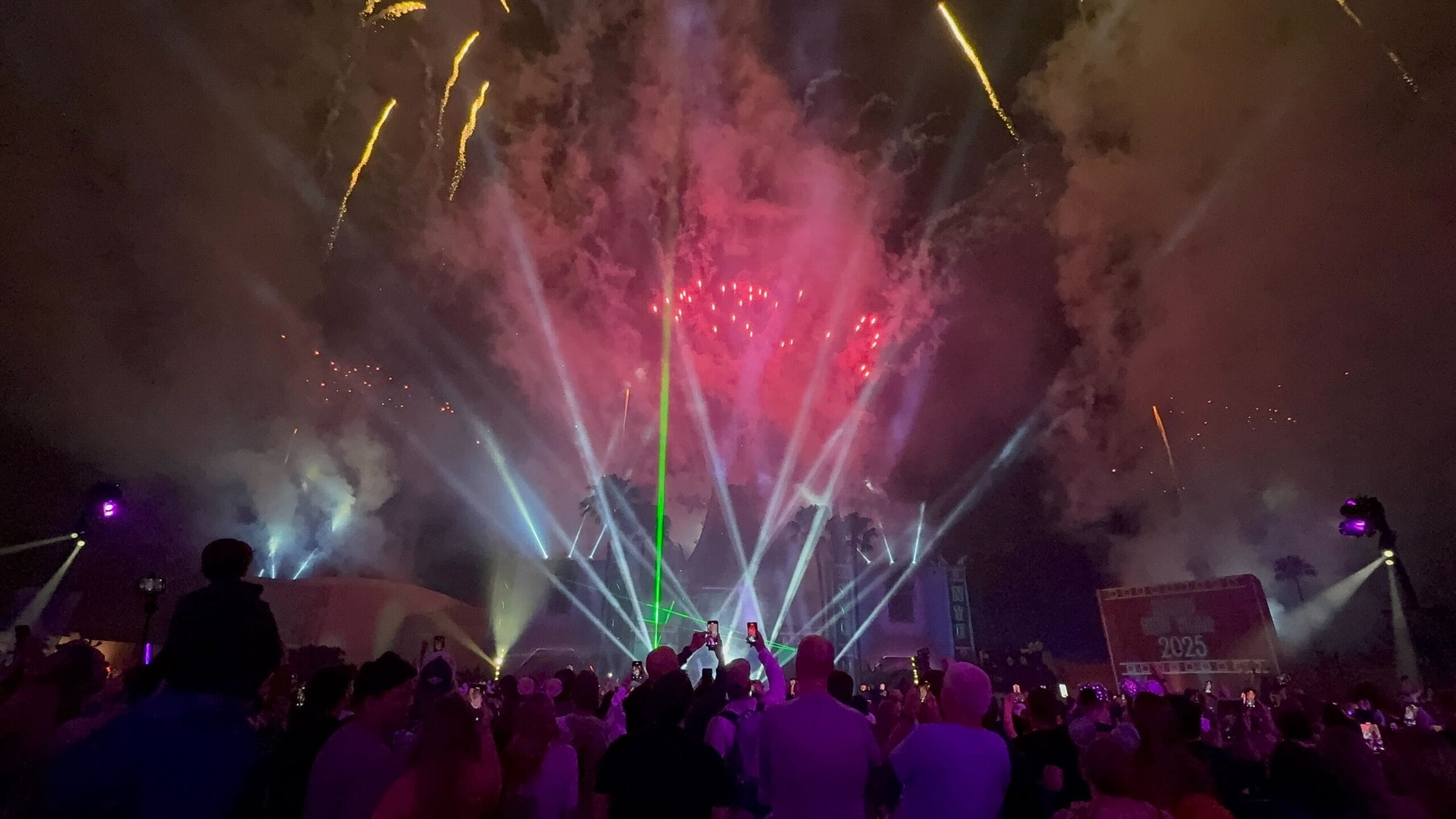 A crowd gathers to watch a dazzling New Year's Eve fireworks display, vibrant colors and smoke painting the night sky.