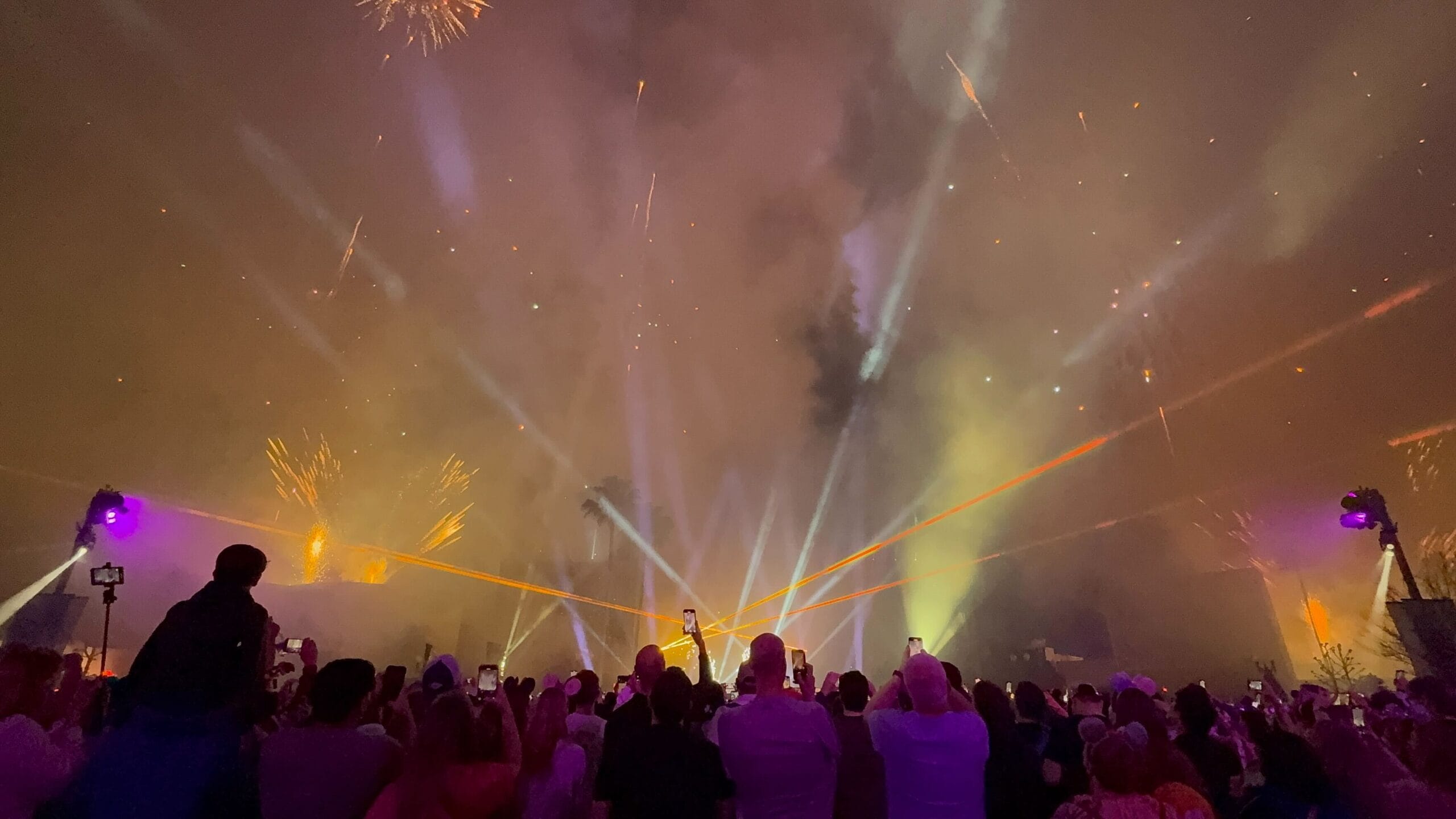 A crowd gathers to watch a vibrant fireworks and laser display light up the New Year's Eve sky, eagerly capturing the dazzling scene with their phones.