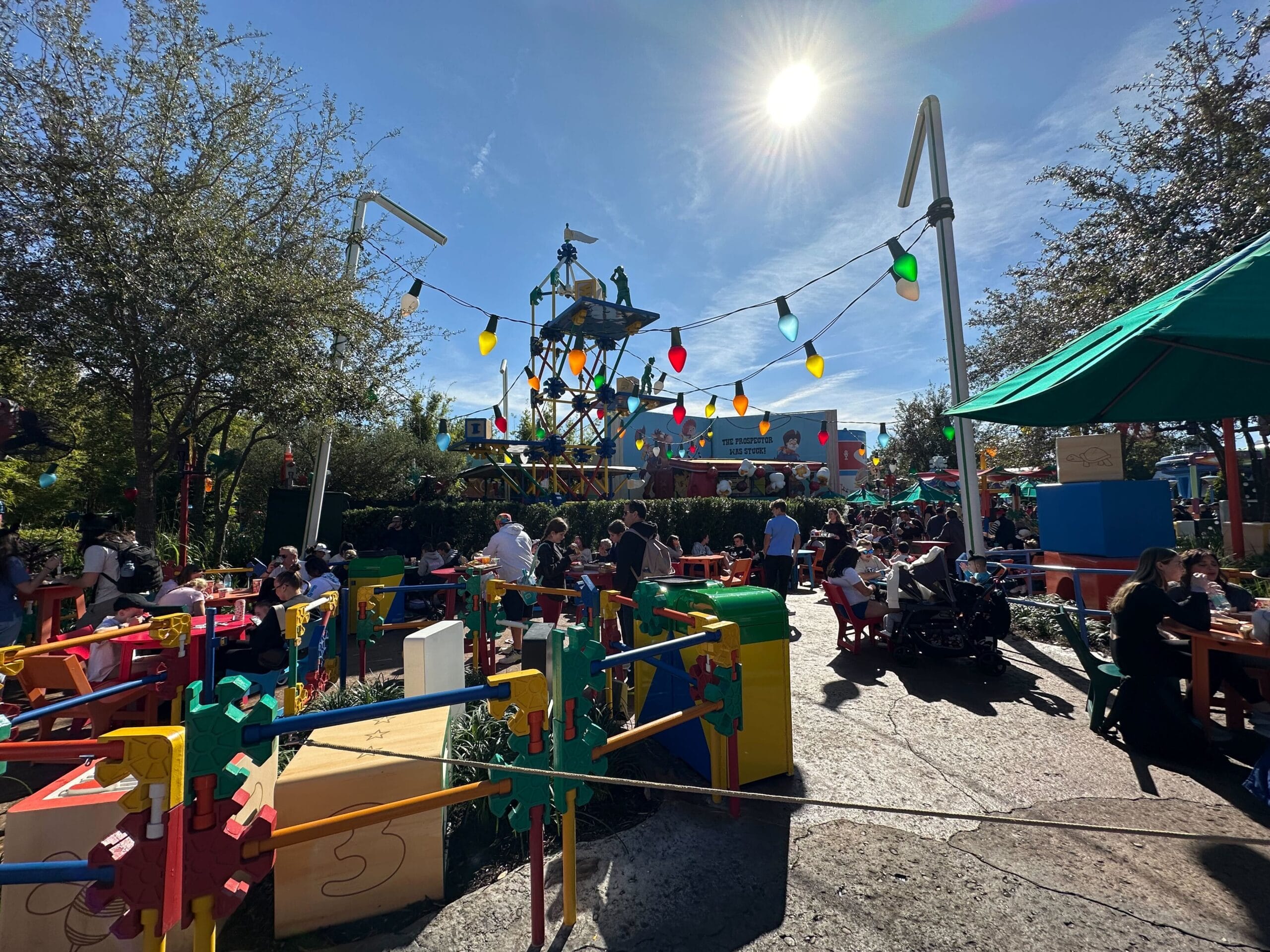 Sunny day at Woody's Lunch Box, an outdoor dining area with colorful decor resembling a child's playset. People are seated under umbrellas and covered seating options, enjoying their meals in the vibrant atmosphere.