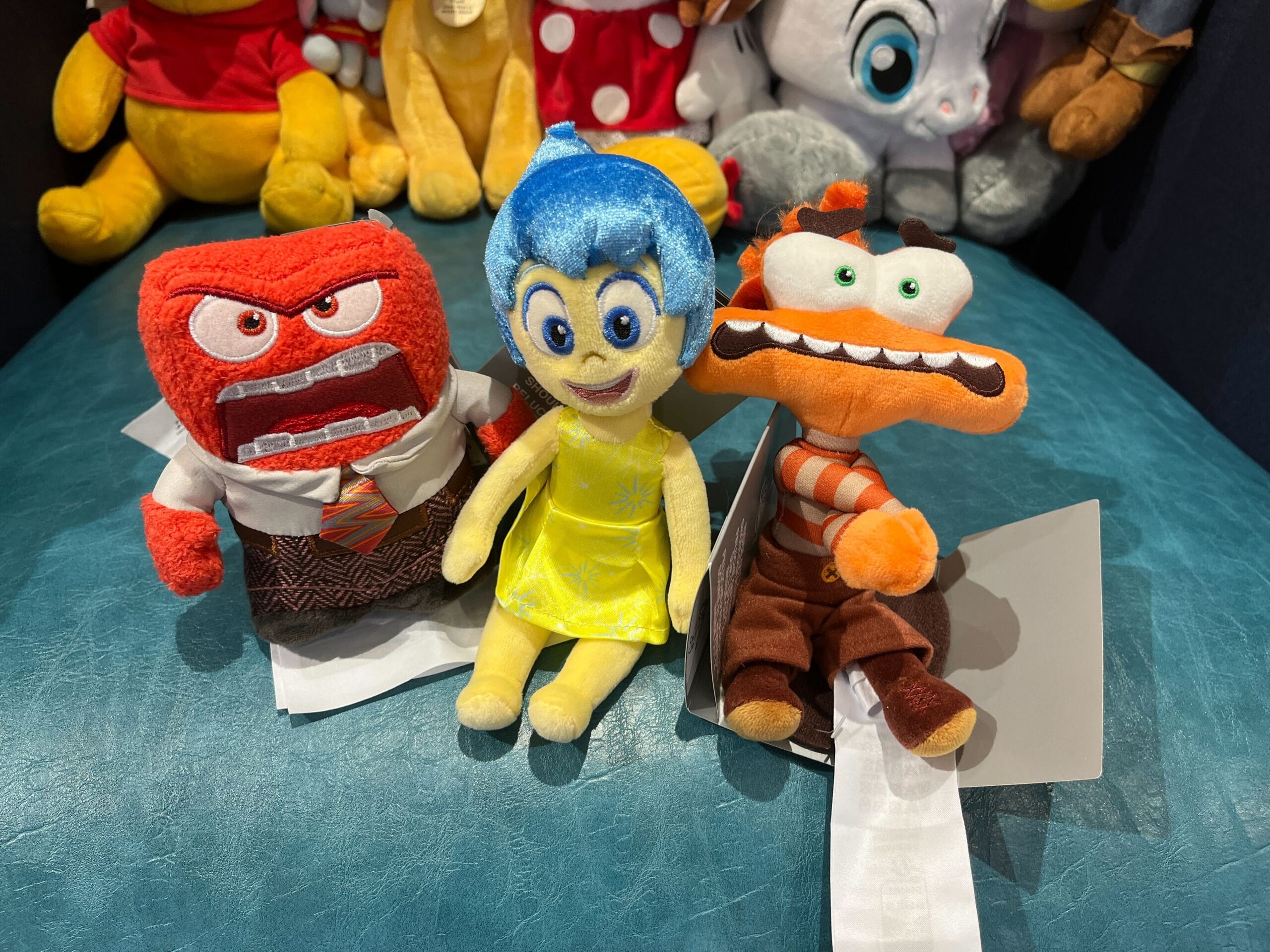Three plush toys with distinct facial expressions are seated on a blue surface, surrounded by other stuffed animals in the background.