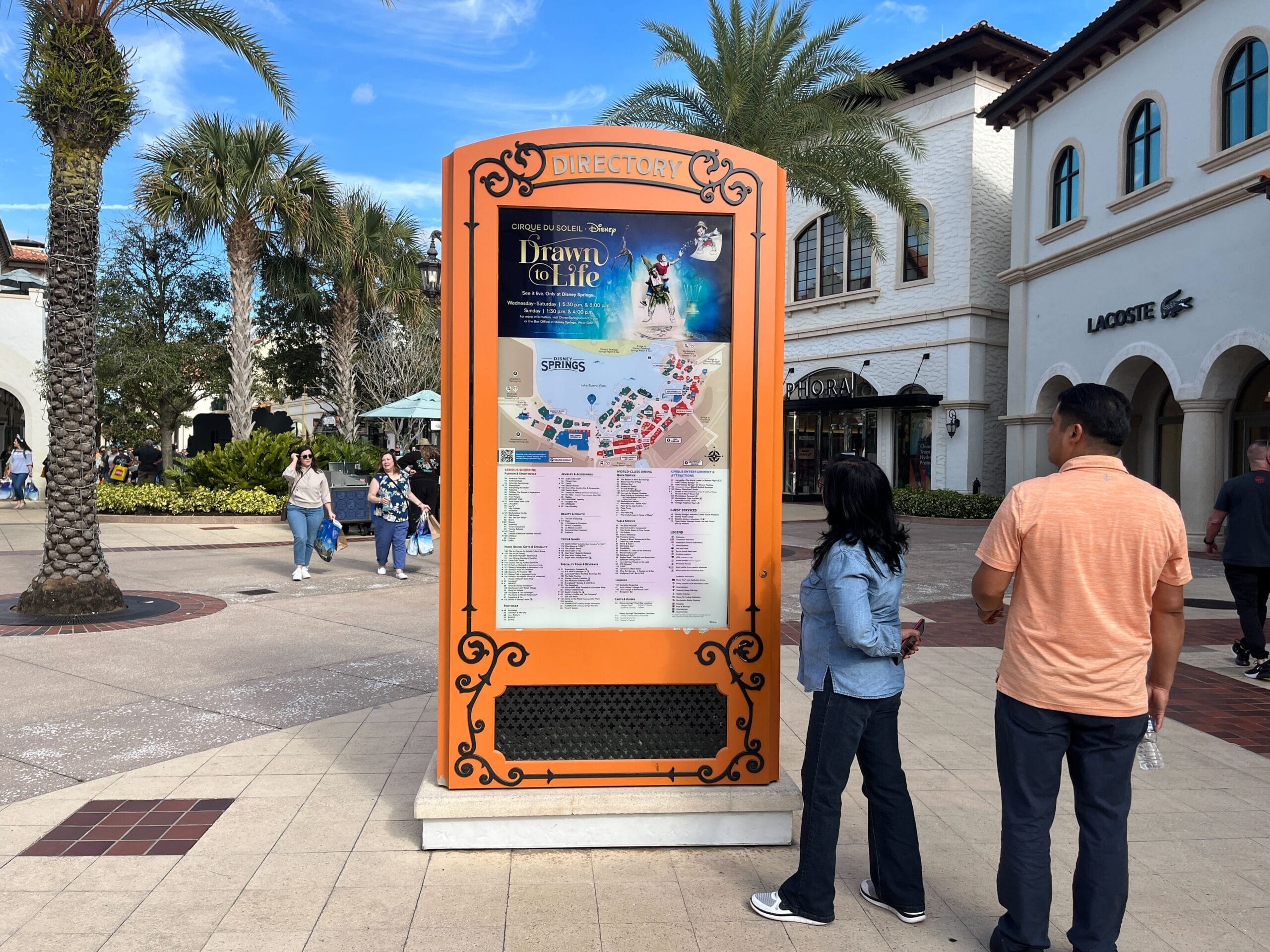 Two people stand in front of an outdoor directory kiosk with a map and event information. Palm trees and stores are visible in the background.
