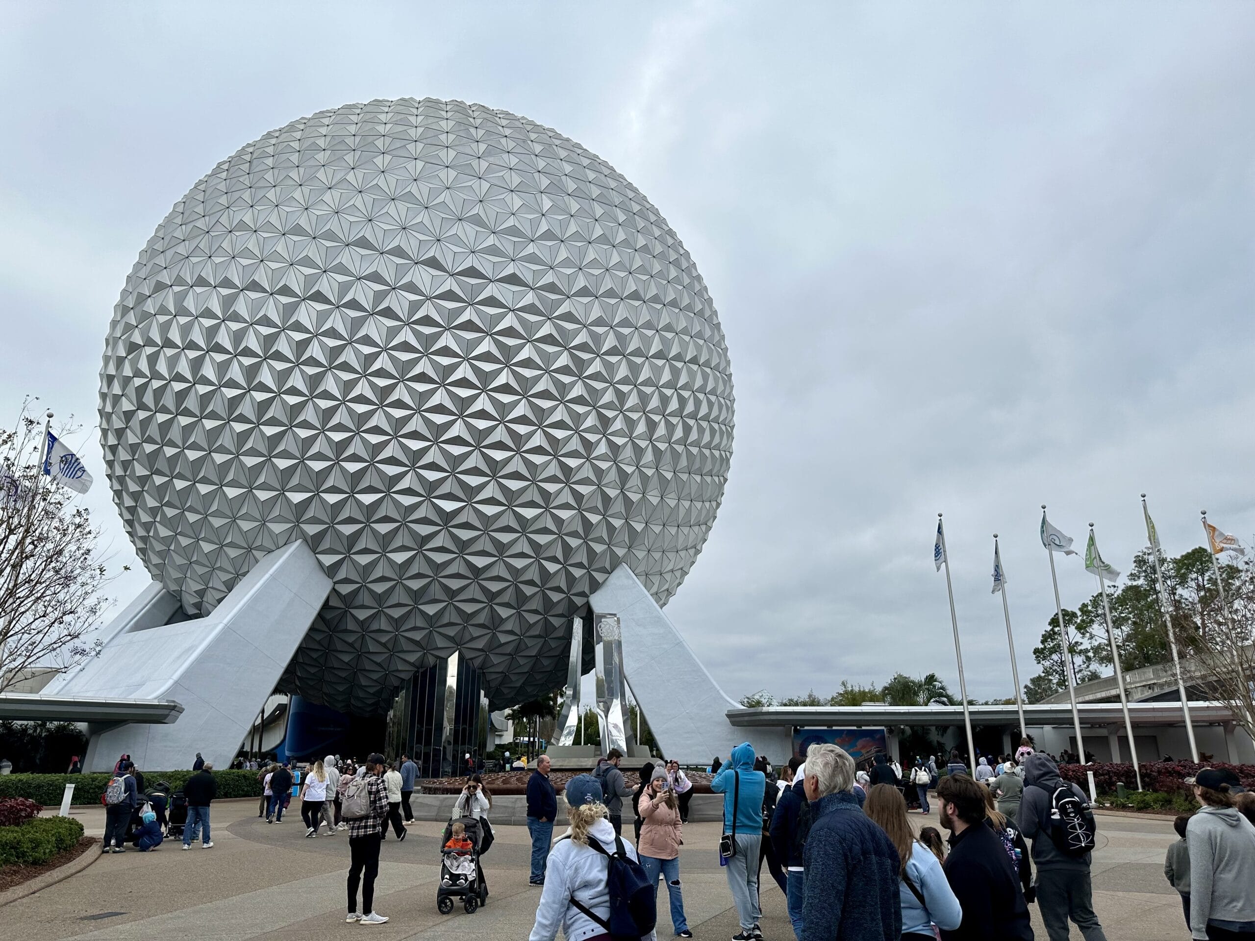 People stand outside the large geodesic sphere of Spaceship Earth at Epcot, surrounded by trees and flags against a cloudy sky.