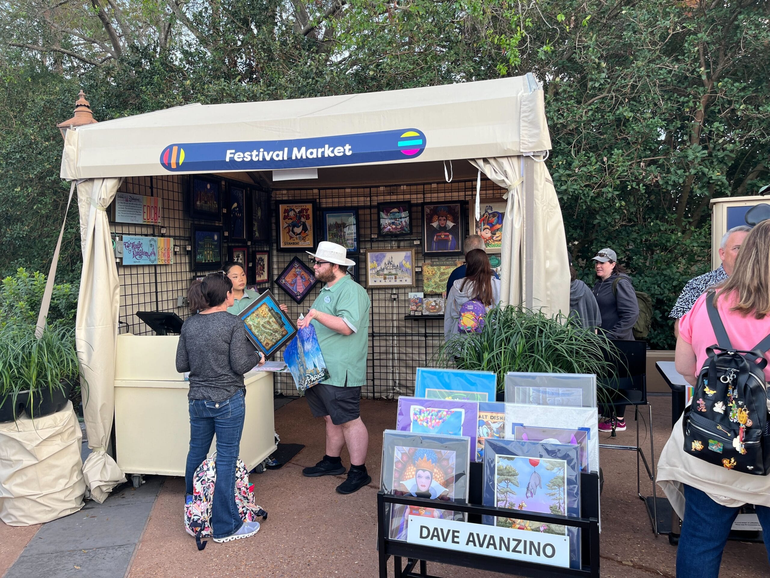 Amid the vibrant atmosphere of the 2025 EPCOT Festival of the Arts, people stroll through a bustling booth adorned with framed pictures. A man in a hat eagerly examines a piece by Dave Avanzino, chatting animatedly with the seller beneath "Festival Market" signs.