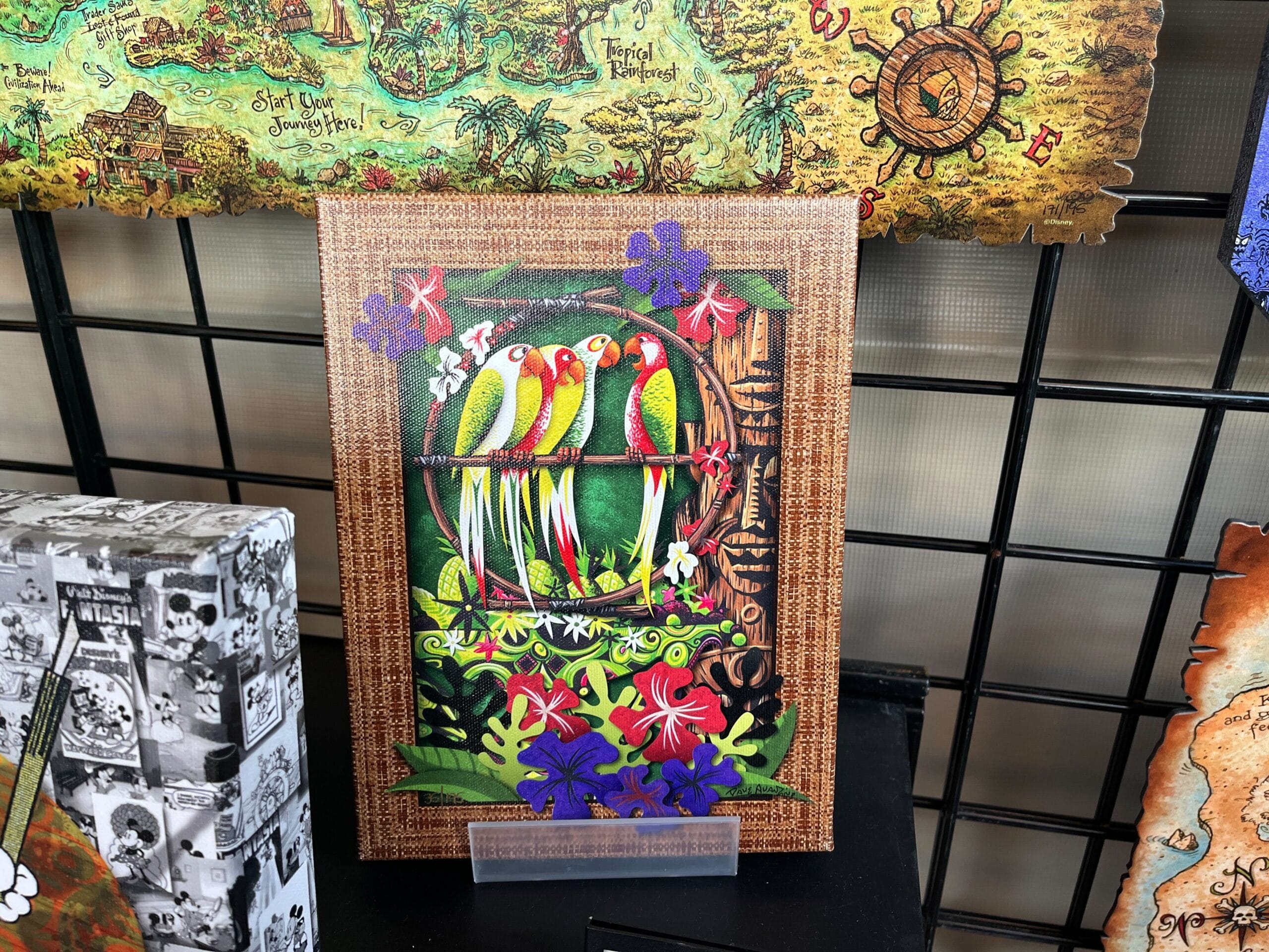 Vibrant artwork by Dave Avazano showcases birds perched on a branch amidst lush flowers, beautifully displayed on a dark grid wall. The piece captures the enchanting spirit of EPCOT's Festival of the Arts.