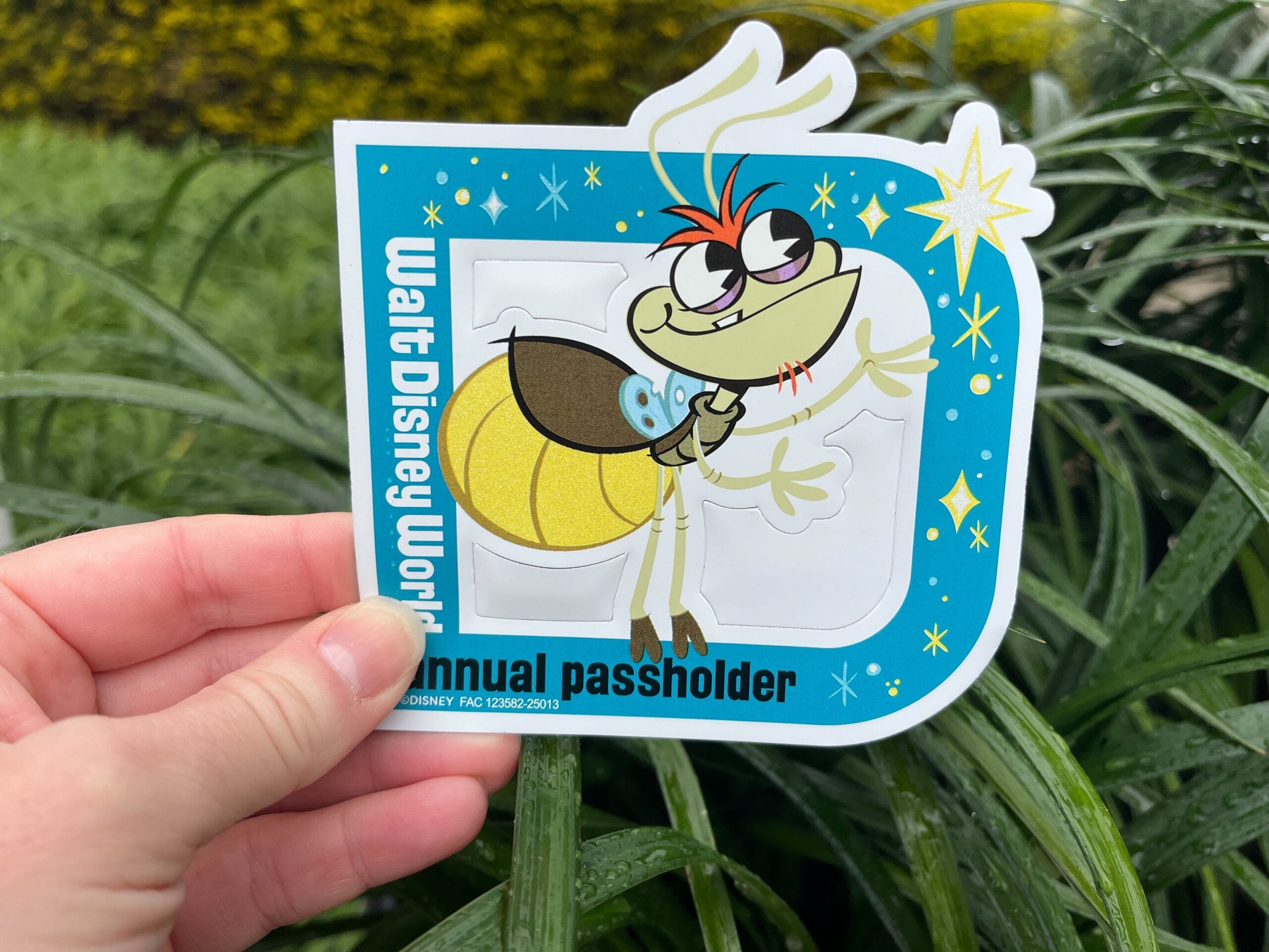 A hand holds a Walt Disney World annual passholder sticker featuring a cartoon bug character against a background of green plants.