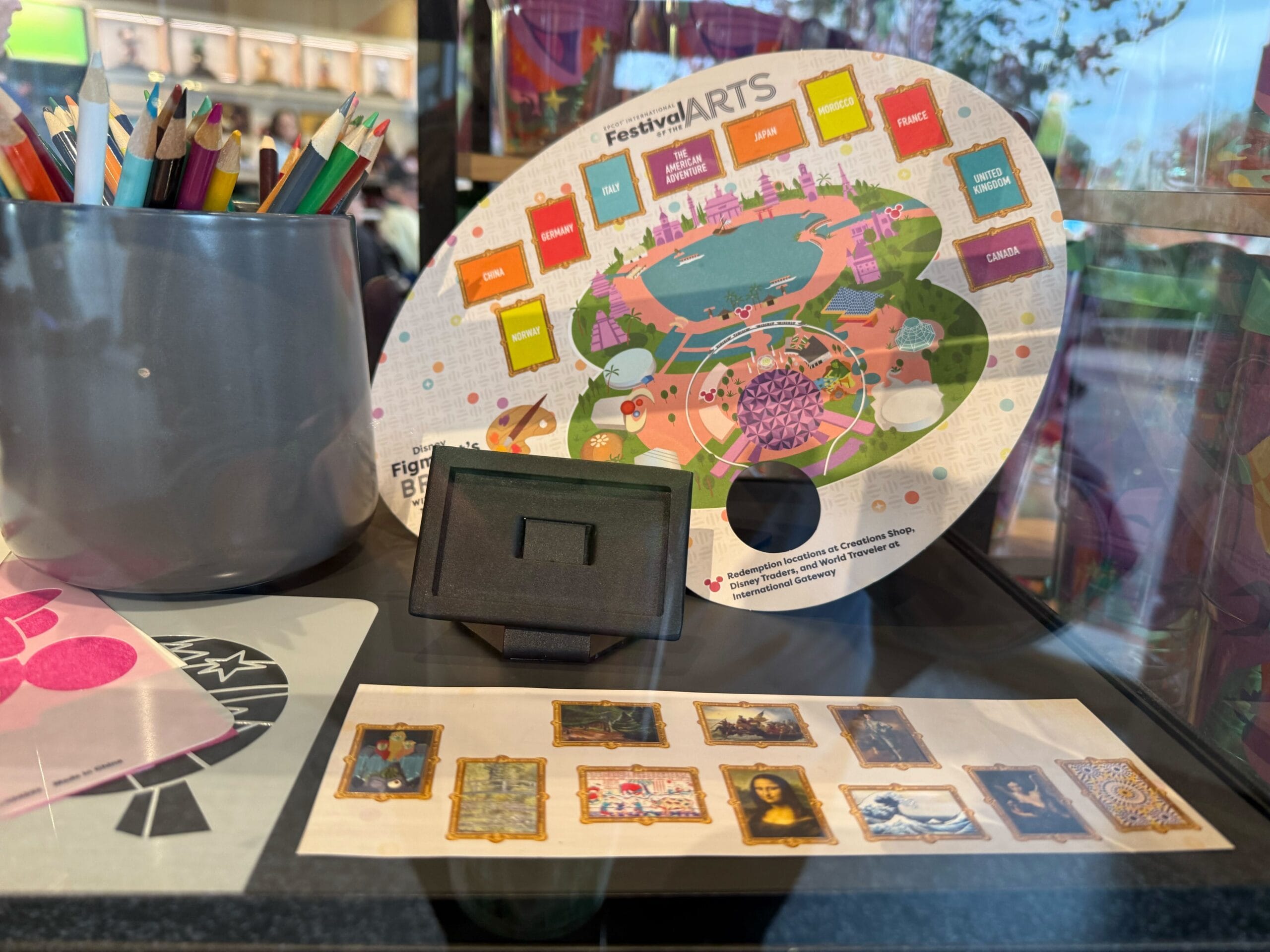 A colorful, art-themed display features a paint palette with labeled sections and colored pencils in a cup, reminiscent of Figment's Brush with the Masters Scavenger Hunt. A collection of small picture frames adorns the countertop, adding to the creative charm.