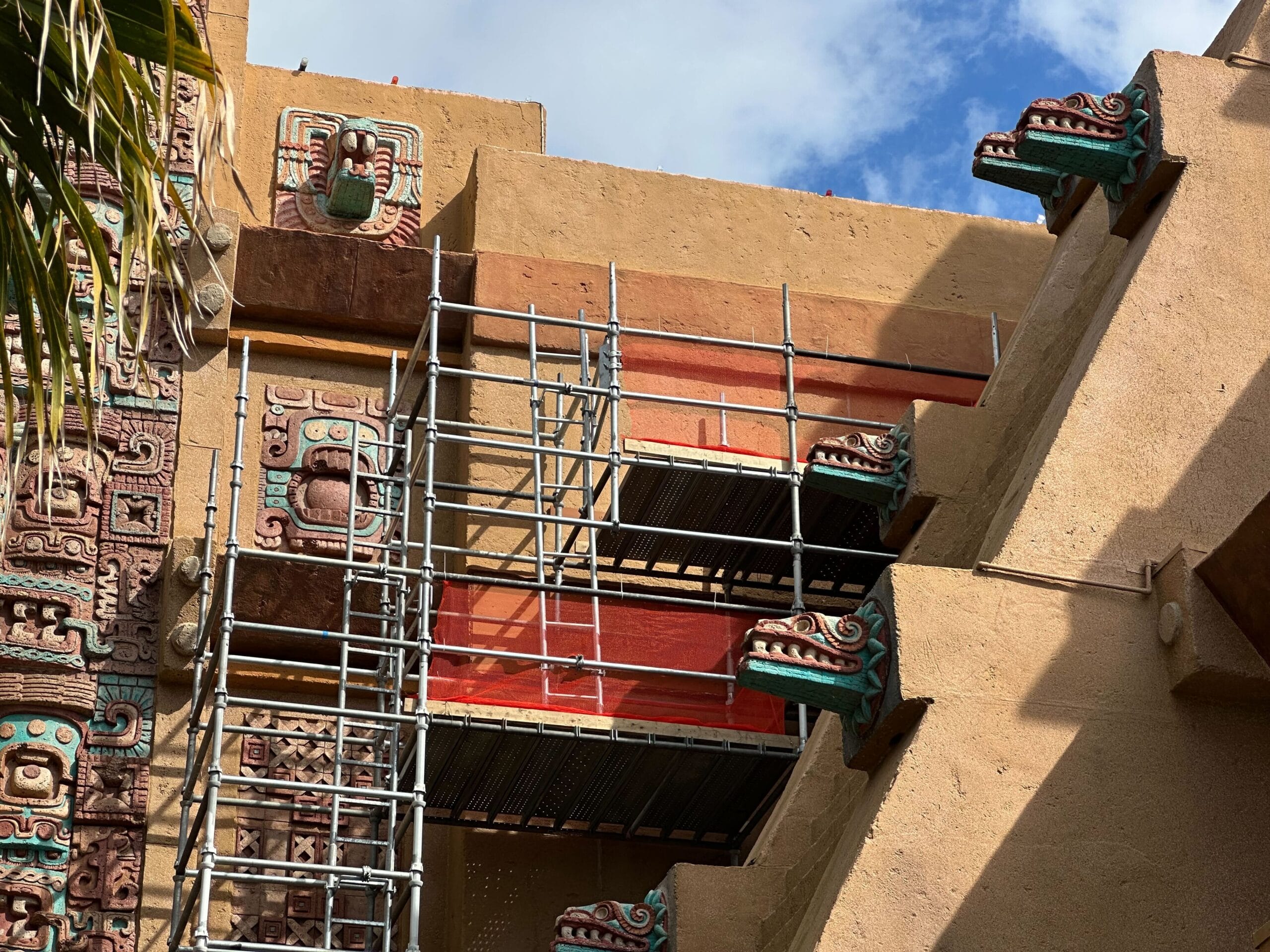 A scaffolding is set up on the beige, carved architectural structure of the Mexico Pavilion at EPCOT, showcasing its decorative elements against a blue sky as the building undergoes repainting.