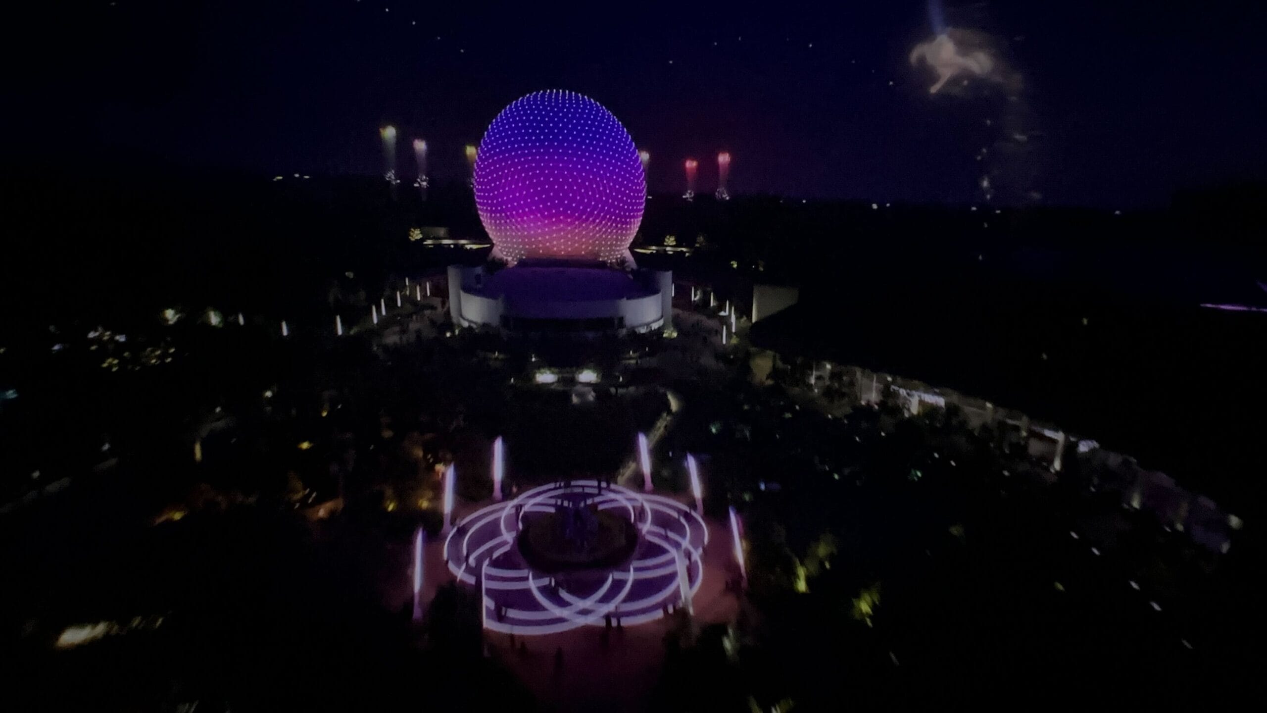 EPCOT’s Spaceship Earth glows purple at night at Disney World, with fountains, lights, and fireworks in the sky.