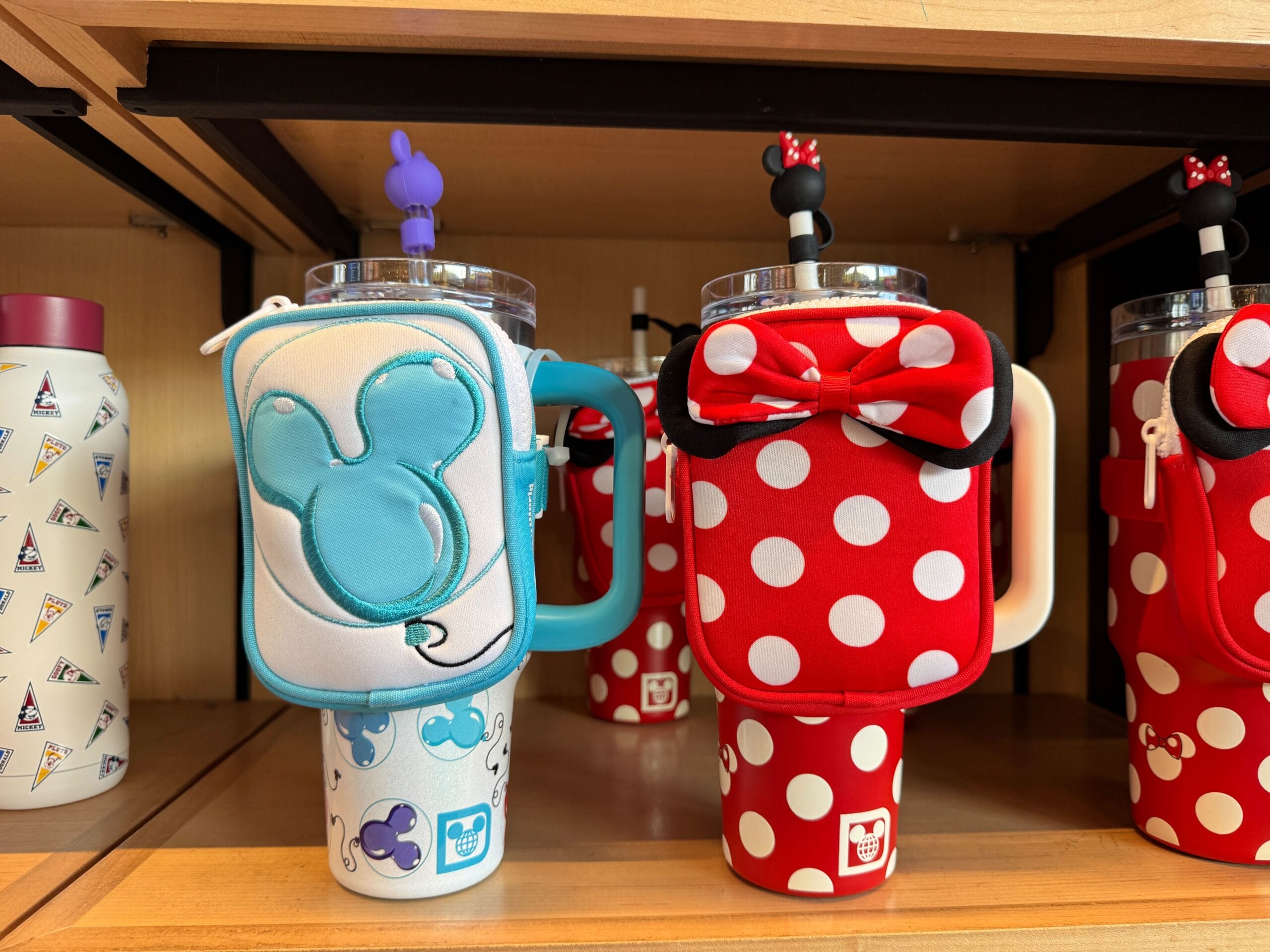 Two themed tumblers with character motifs and covers, resembling Stanley-Style Minnie Mouse under the magic of Walt Disney World. One boasts a blue design, while the other is red with white polka dots and a bow. They make a charming display on a wooden shelf.