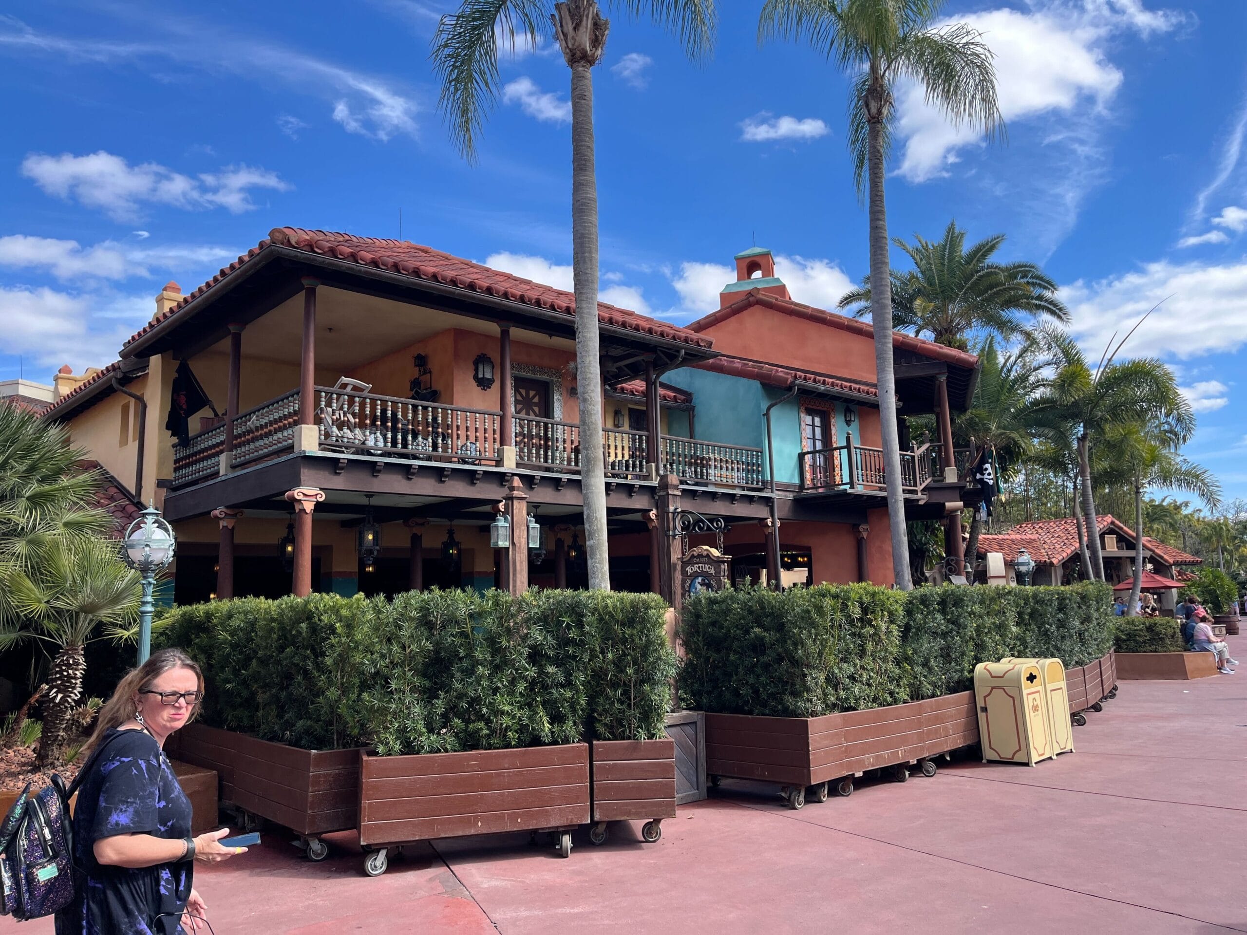 A two-story building with a balcony and brown railing, flanked by palm trees. A woman walks by with a backpack under a clear blue sky.