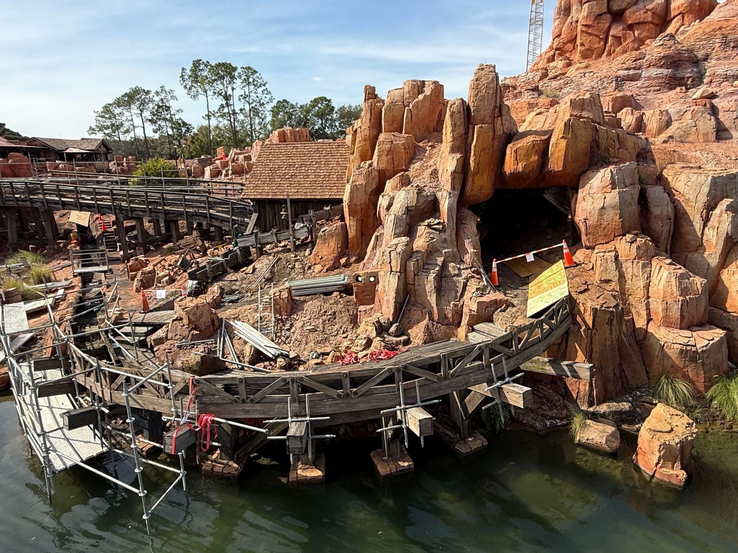 Amusement park roller coaster under maintenance, with exposed tracks and scaffolding near a rocky structure by water.