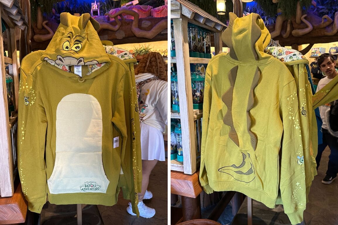 The yellow dragon-themed cosplay hoodie features cartoonish eyes and teeth on the hood, a white belly, and a tail design on the back. Displayed hanging in a store with other merchandise visible, it’s perfect for your next adventure at Walt Disney World.