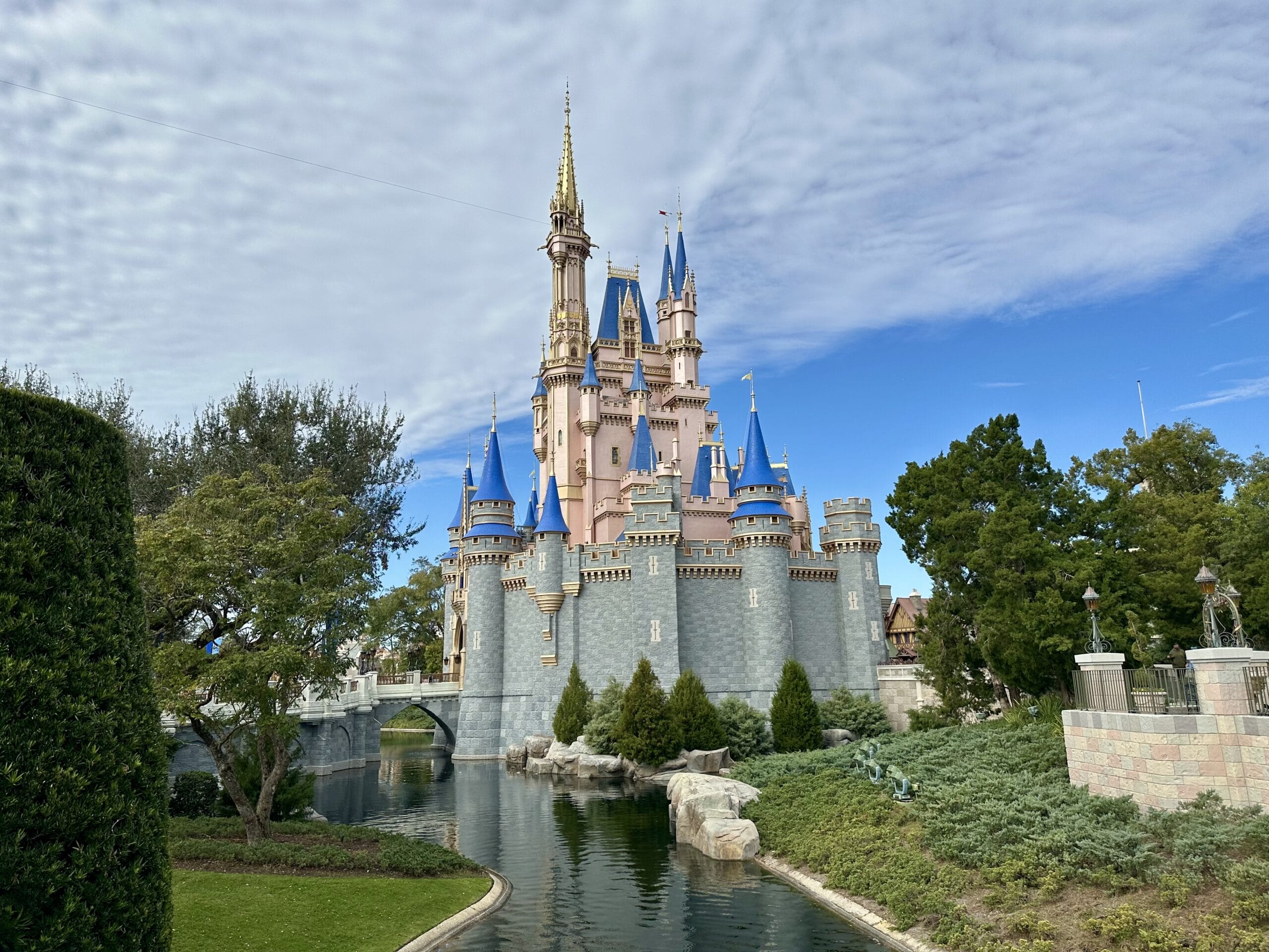 A fairytale castle with blue and gold spires is surrounded by a moat, green trees, and a cloudy sky.