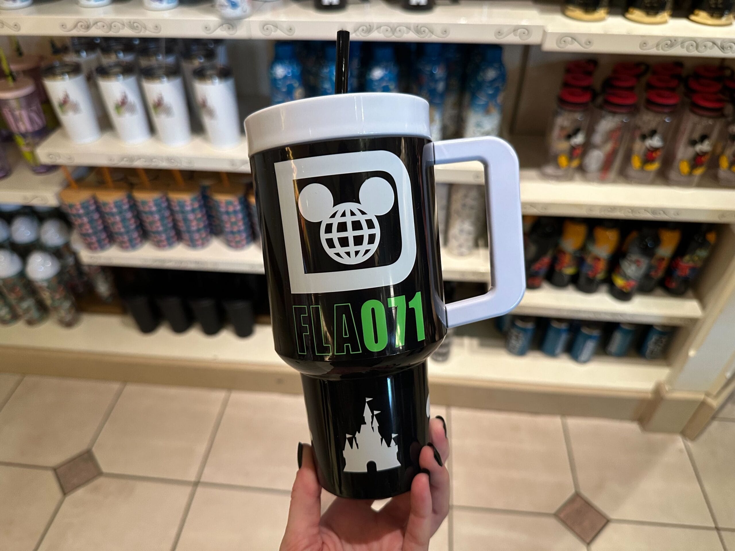A hand holds a black travel mug with a white castle silhouette and green lettering. It has a white handle and a straw. Shelves of similar mugs are in the background.