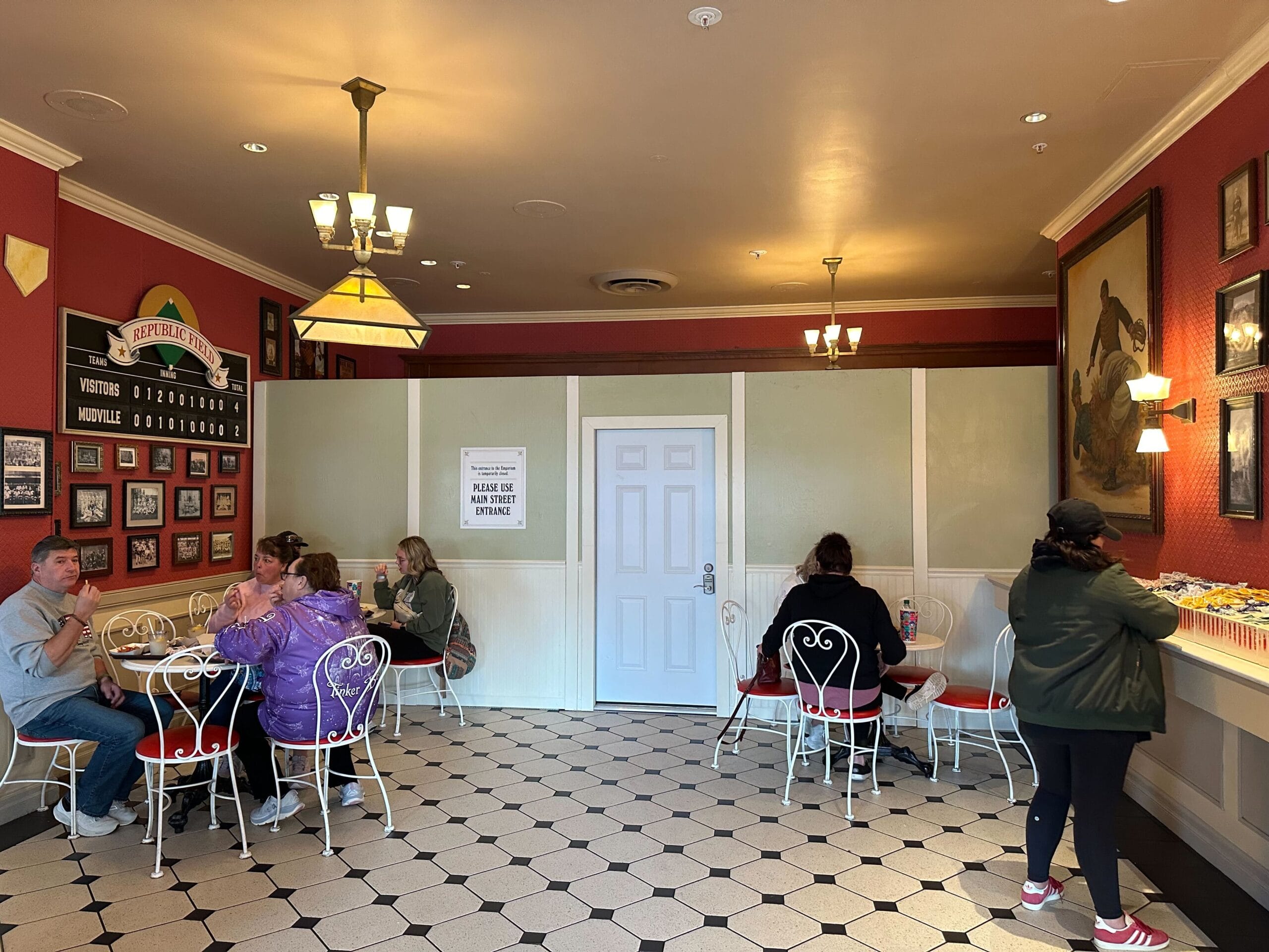 People seated at small tables in a room with red walls and black-and-white floor tiles. A sign on the wall reads, "Please place bags on your lap. Thank you.