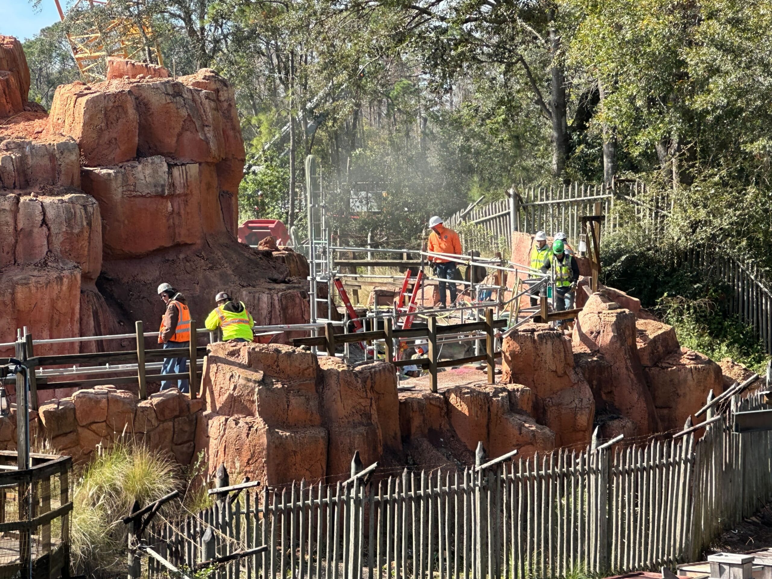Construction workers in safety gear working on a rocky landscape surrounded by wooden fencing, with trees in the background.