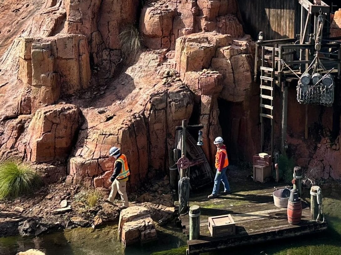 Outdoor scene featuring a rocky, Western-themed structure beside water, with a crane in the background and a few people walking on a path.