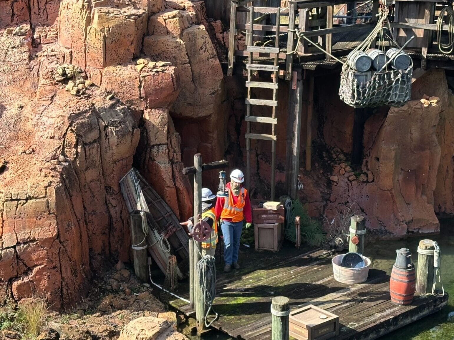A person in a safety vest and helmet stands on a wooden platform near red rock formations and water, next to a ladder and equipment.