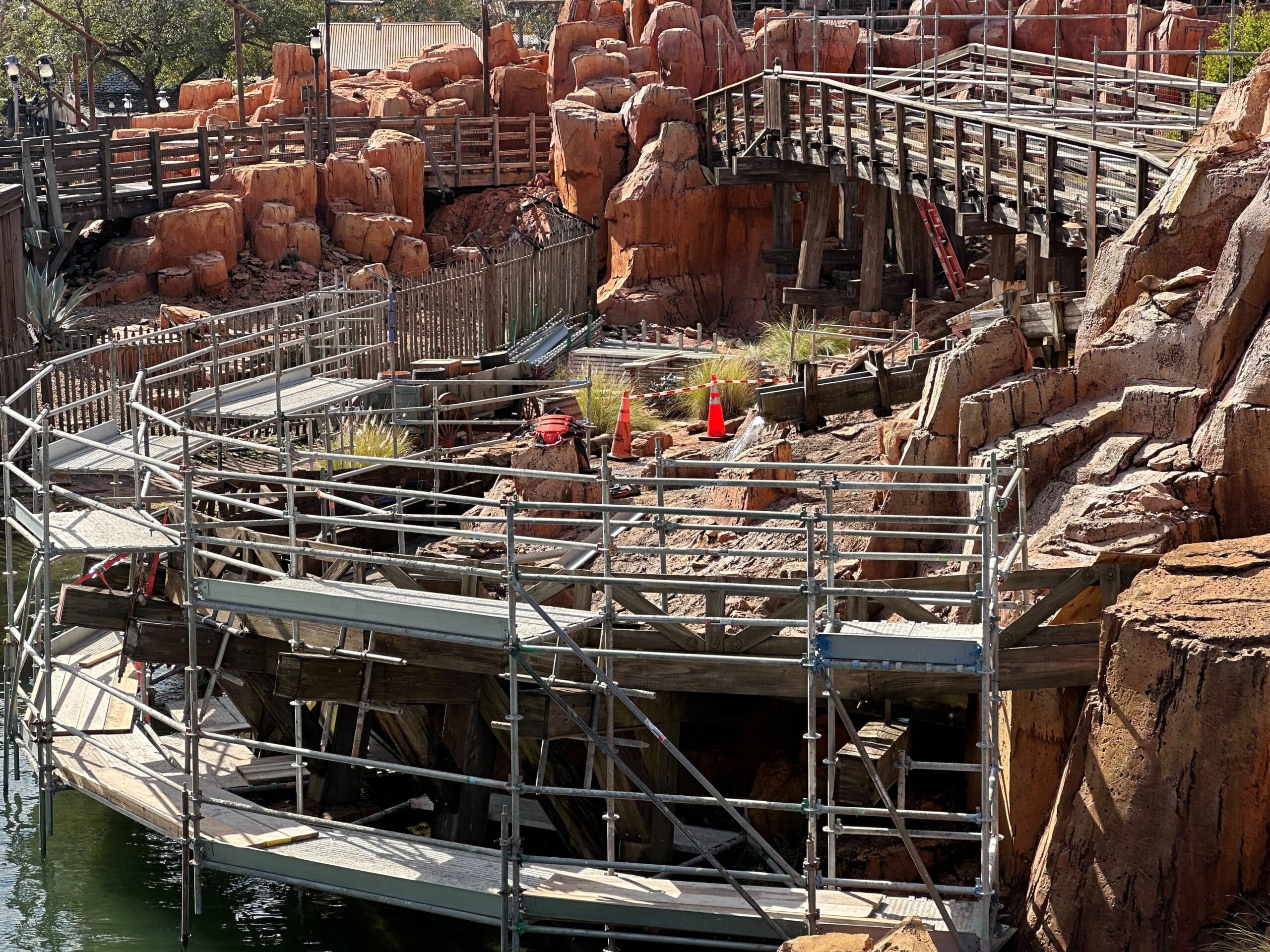 Rocky landscape with wooden walkways and scaffolding, adjacent to a body of water. Orange traffic cones and construction materials are visible amidst the rocky terrain.