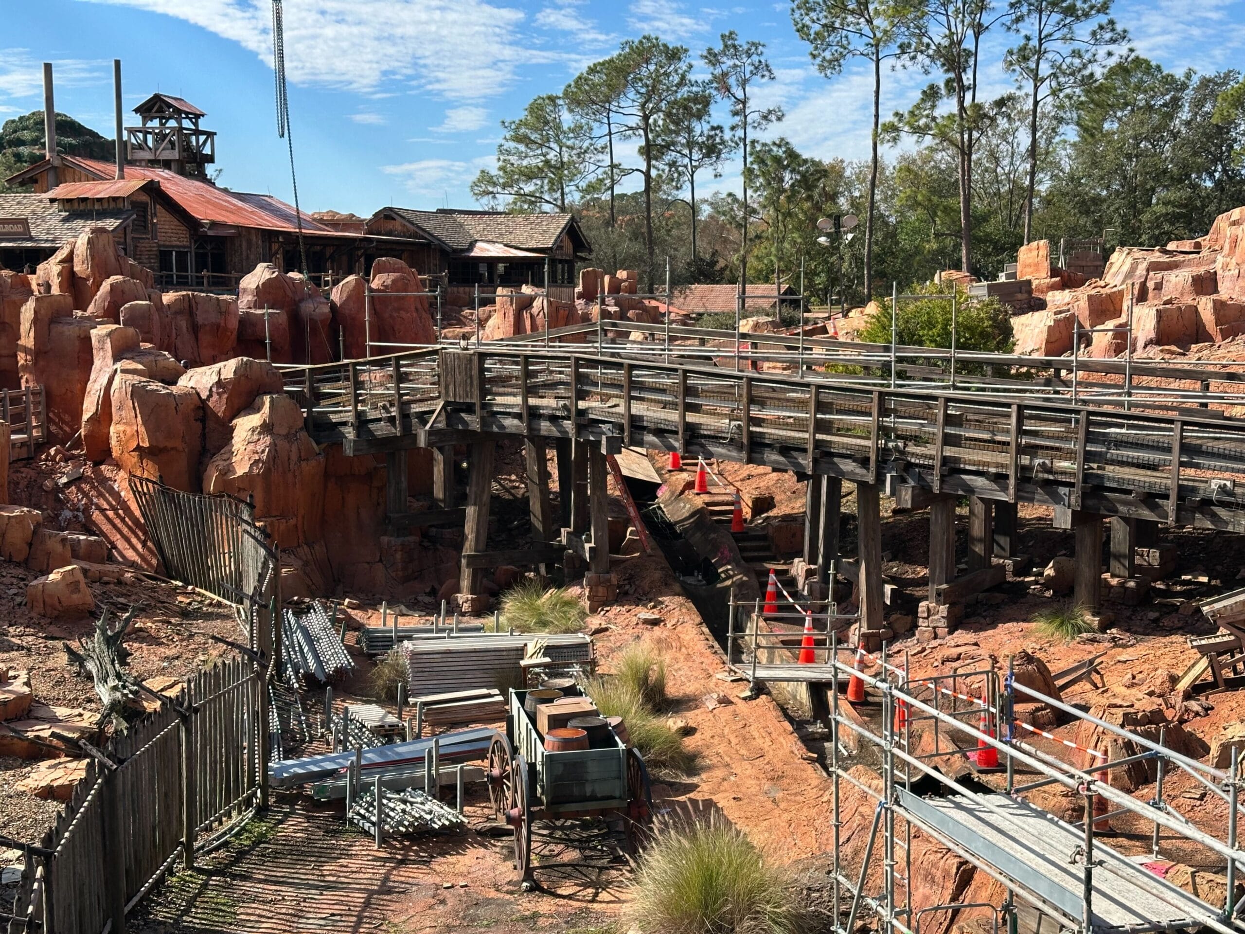 Wooden bridge and rocky terrain under construction with scaffolding and orange cones. Sparse trees in the background, partly cloudy sky.