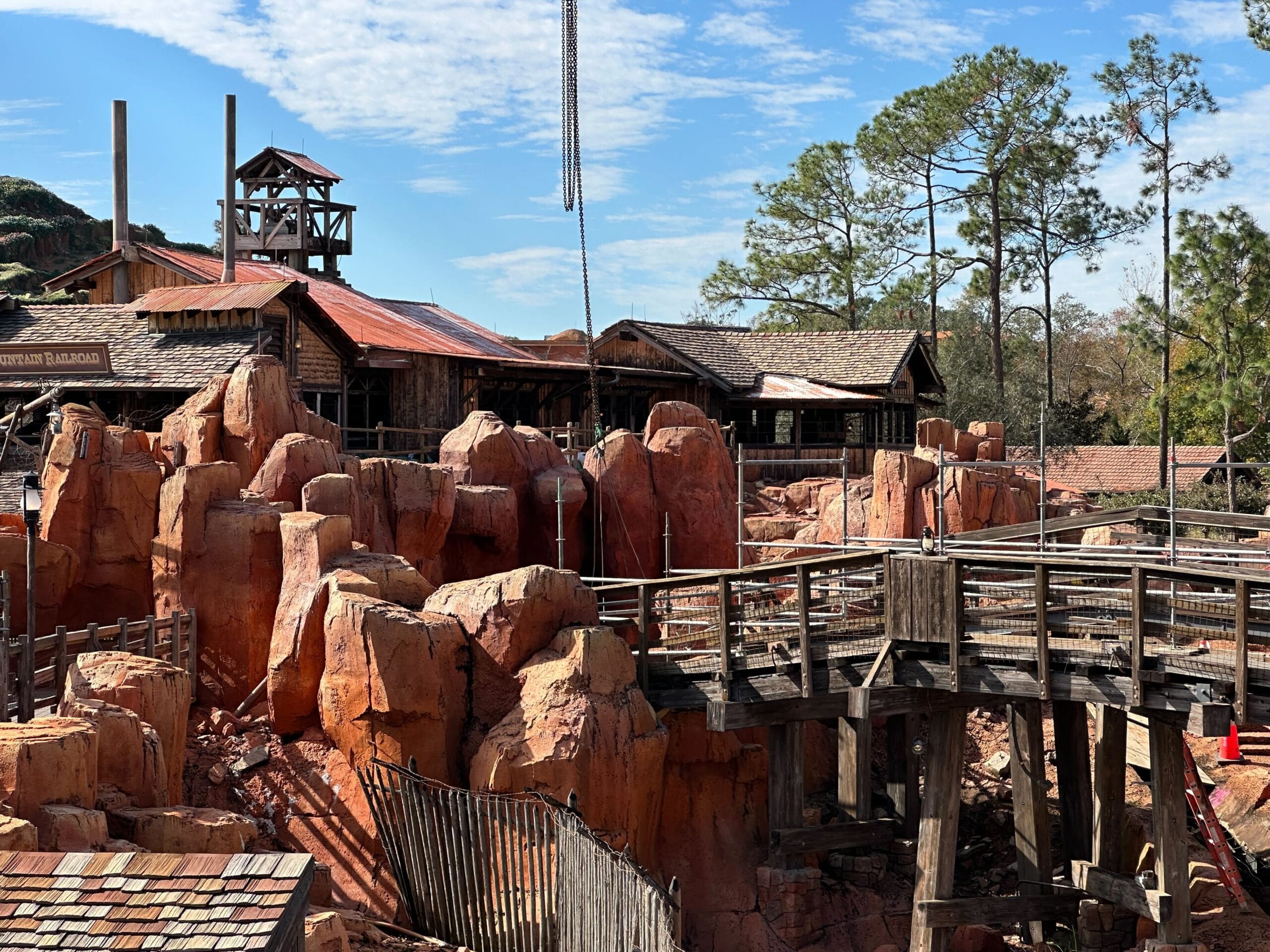 A rustic, wooden-themed amusement park ride with rocky terrain, wooden bridges, and western-style buildings set against a blue sky and trees.