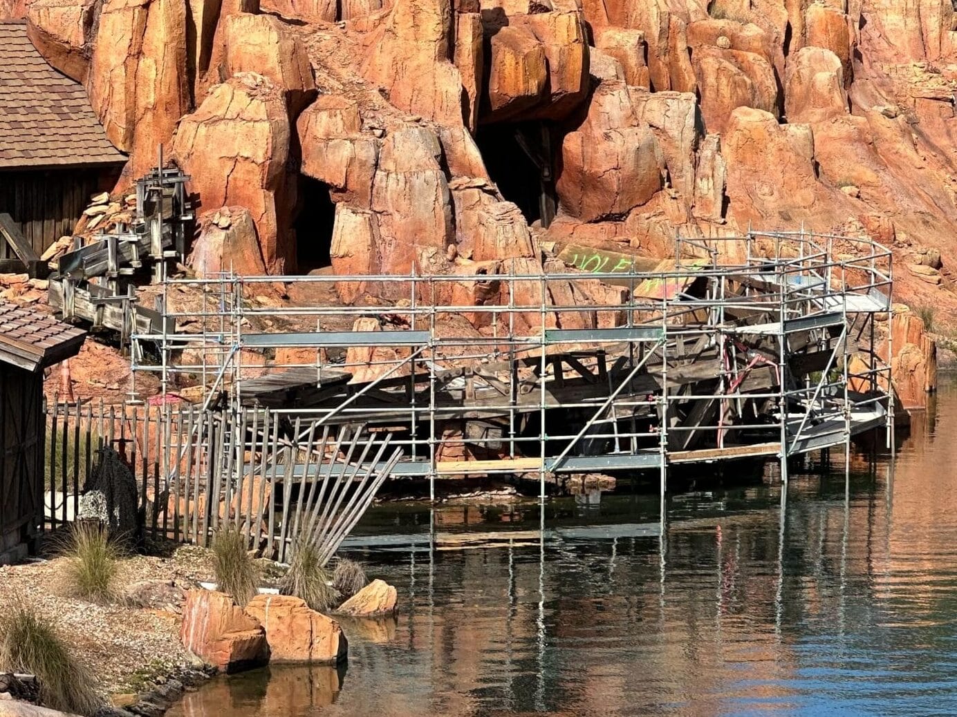 Rocky terrain with wooden structures by a calm body of water. A mountain backdrop and scattered vegetation surround the scene.