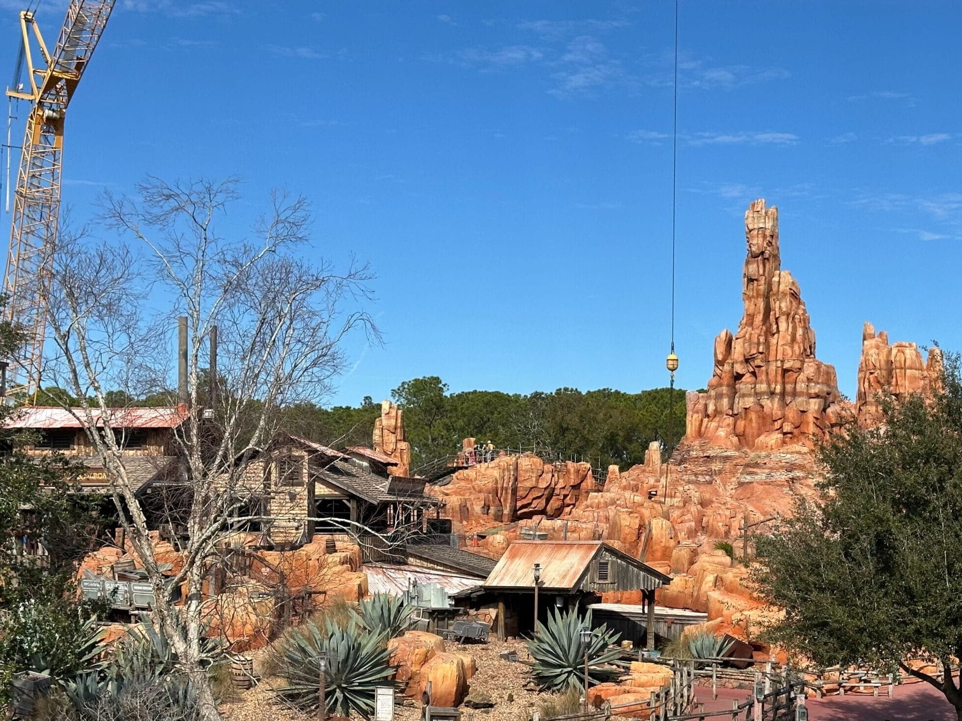 A western-themed amusement park area with rocky formations, wooden structures, desert plants, and a crane in the background under a clear blue sky.