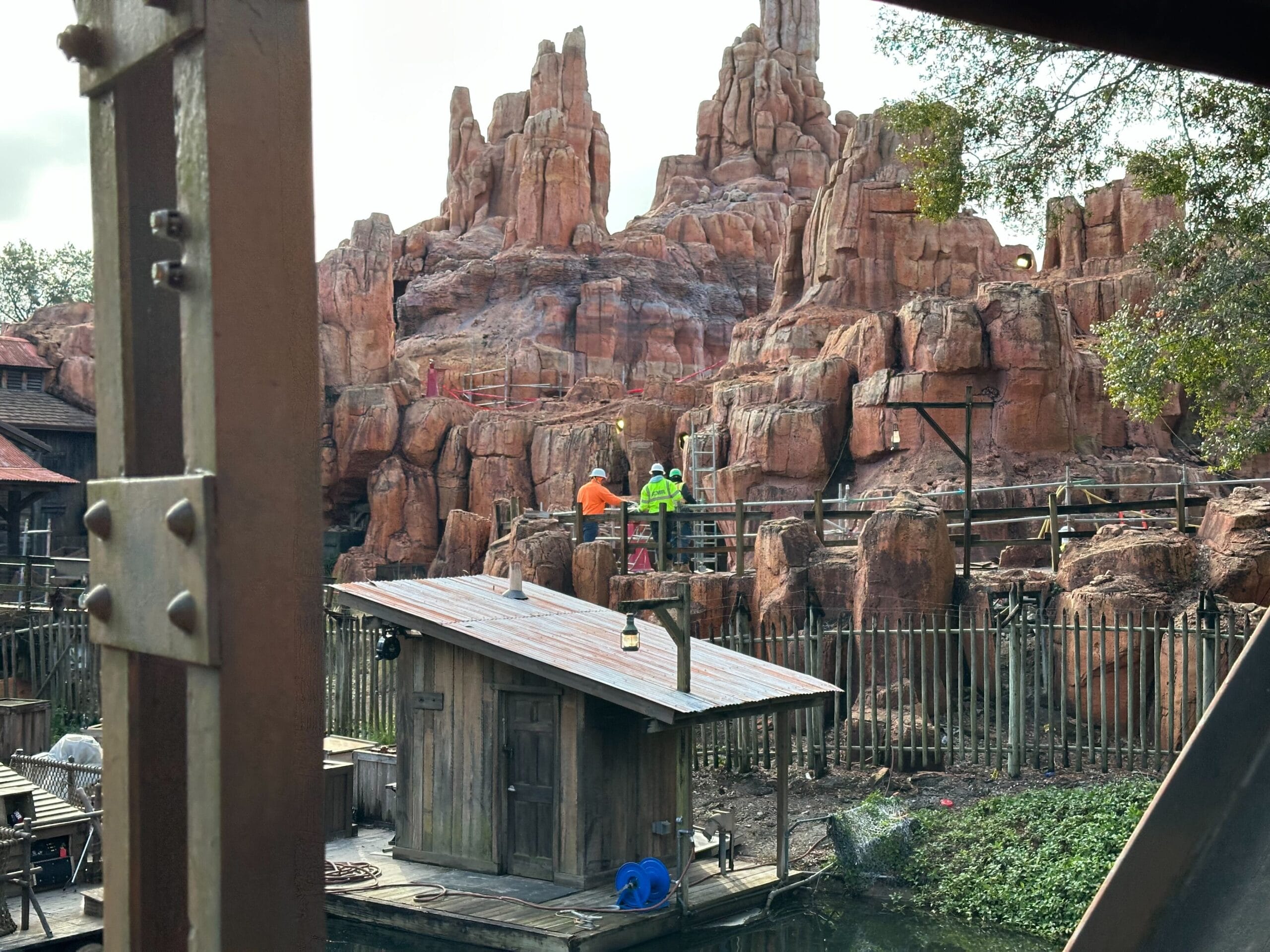 Two people in bright vests work on a wooden platform near a rocky structure at an amusement park. A small building and a body of water are in the foreground.