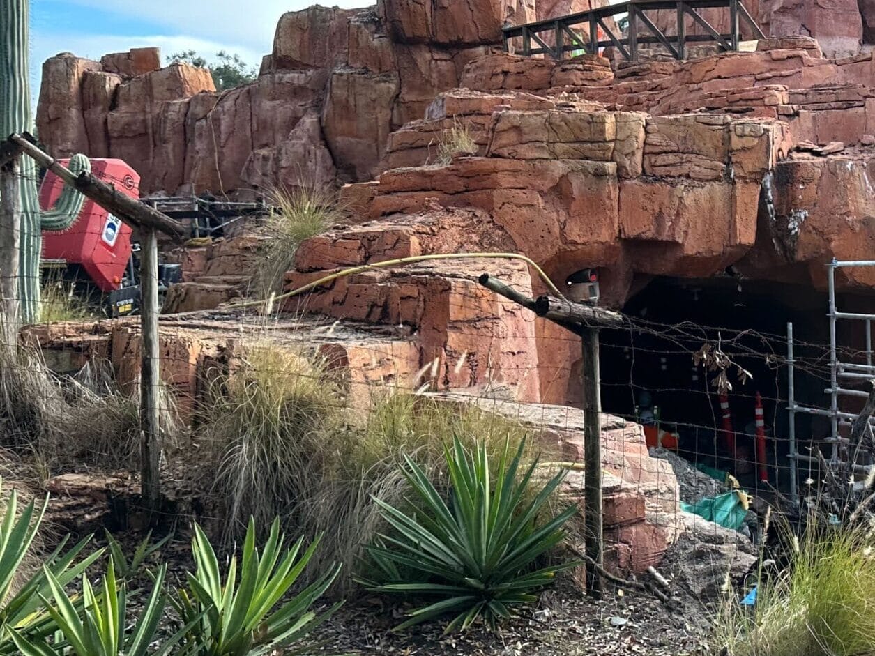 Red rock canyon with wooden structure and path, surrounded by desert plants. Some fencing and a tunnel entrance are visible. Clear sky in the background.