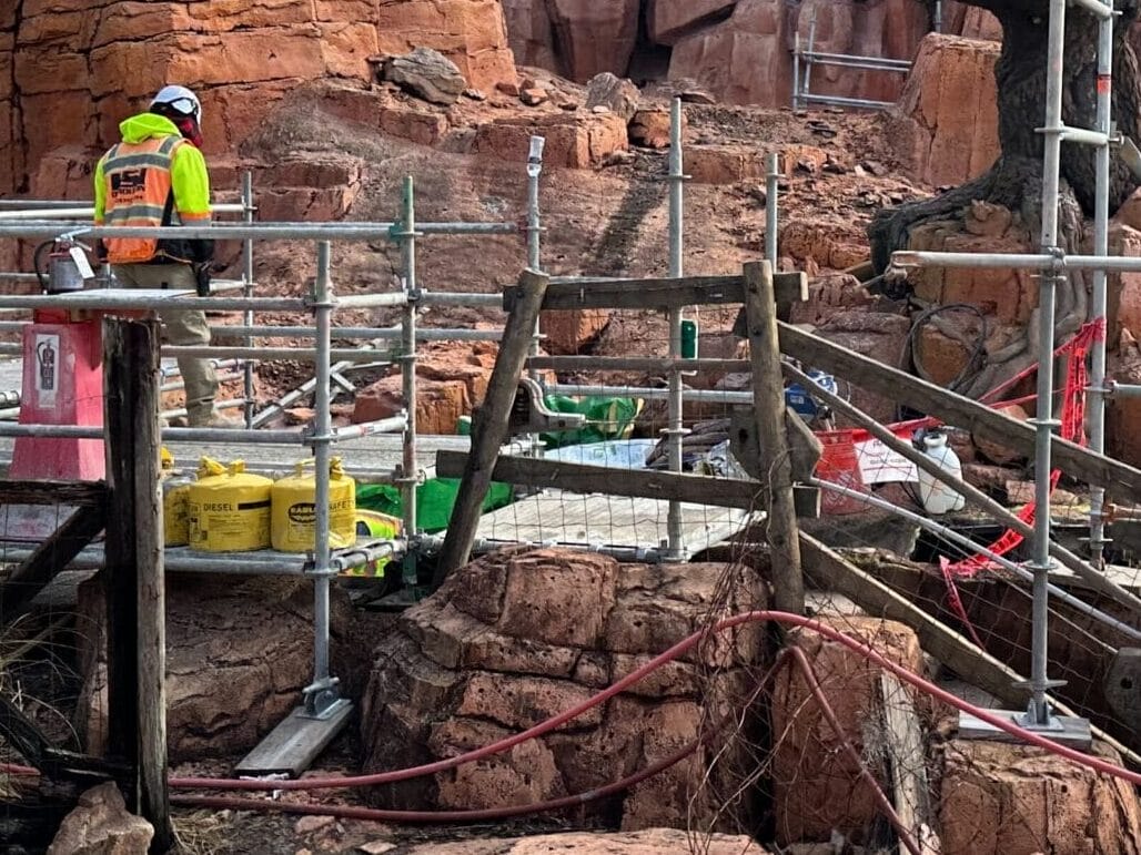 Construction worker stands on a scaffolding amid red rock formations under a blue sky. Wooden railings and construction materials are visible.