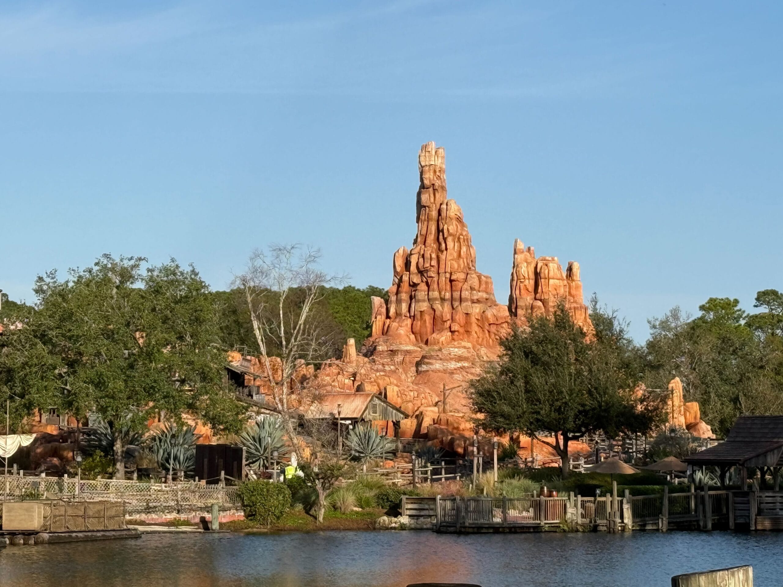 A rocky, red-hued mountain formation set against a clear sky, surrounded by trees and a body of water.