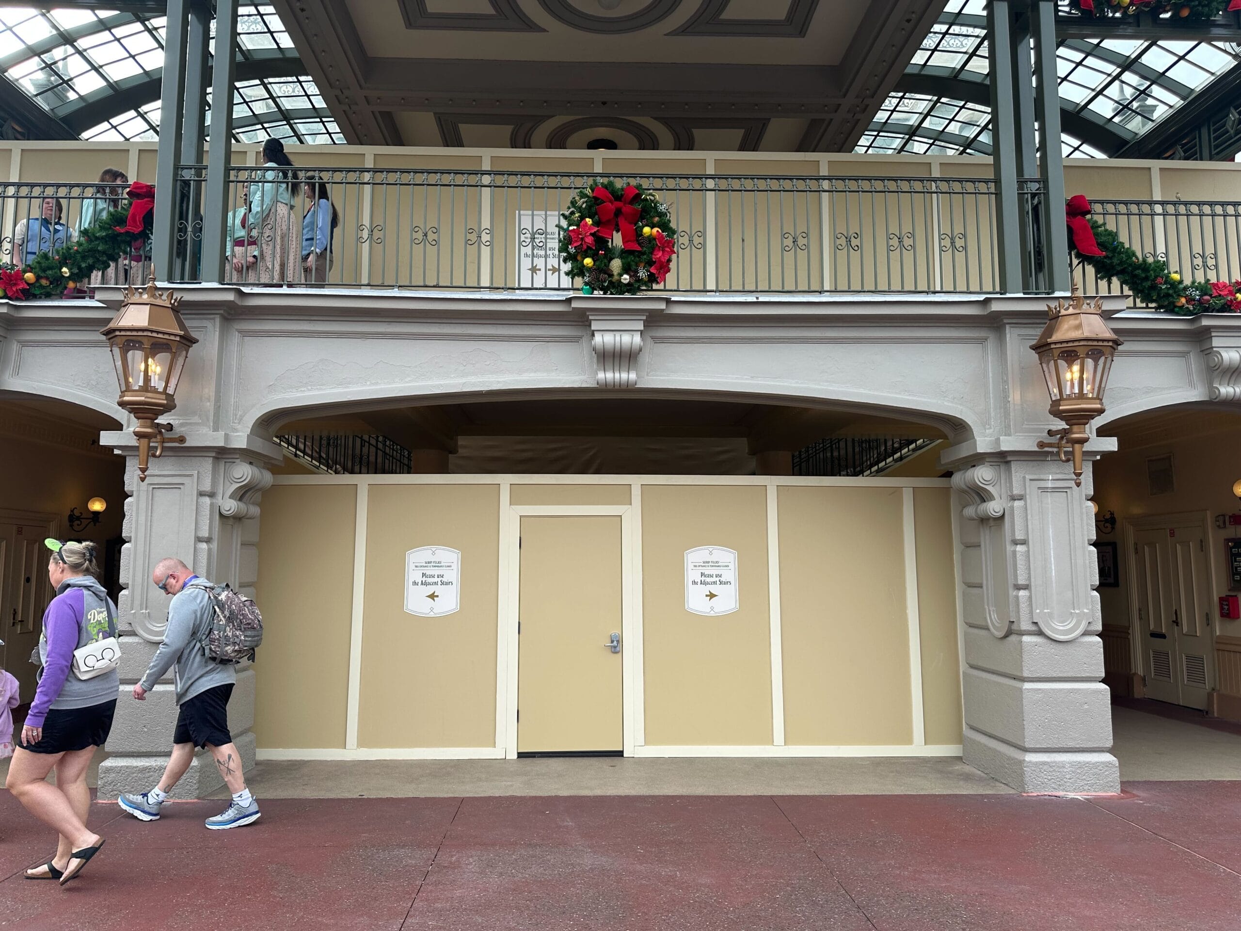 A construction wall with signs blocks an entrance under a balcony. Holiday wreaths adorn the railing above. Two people walk by on the left.