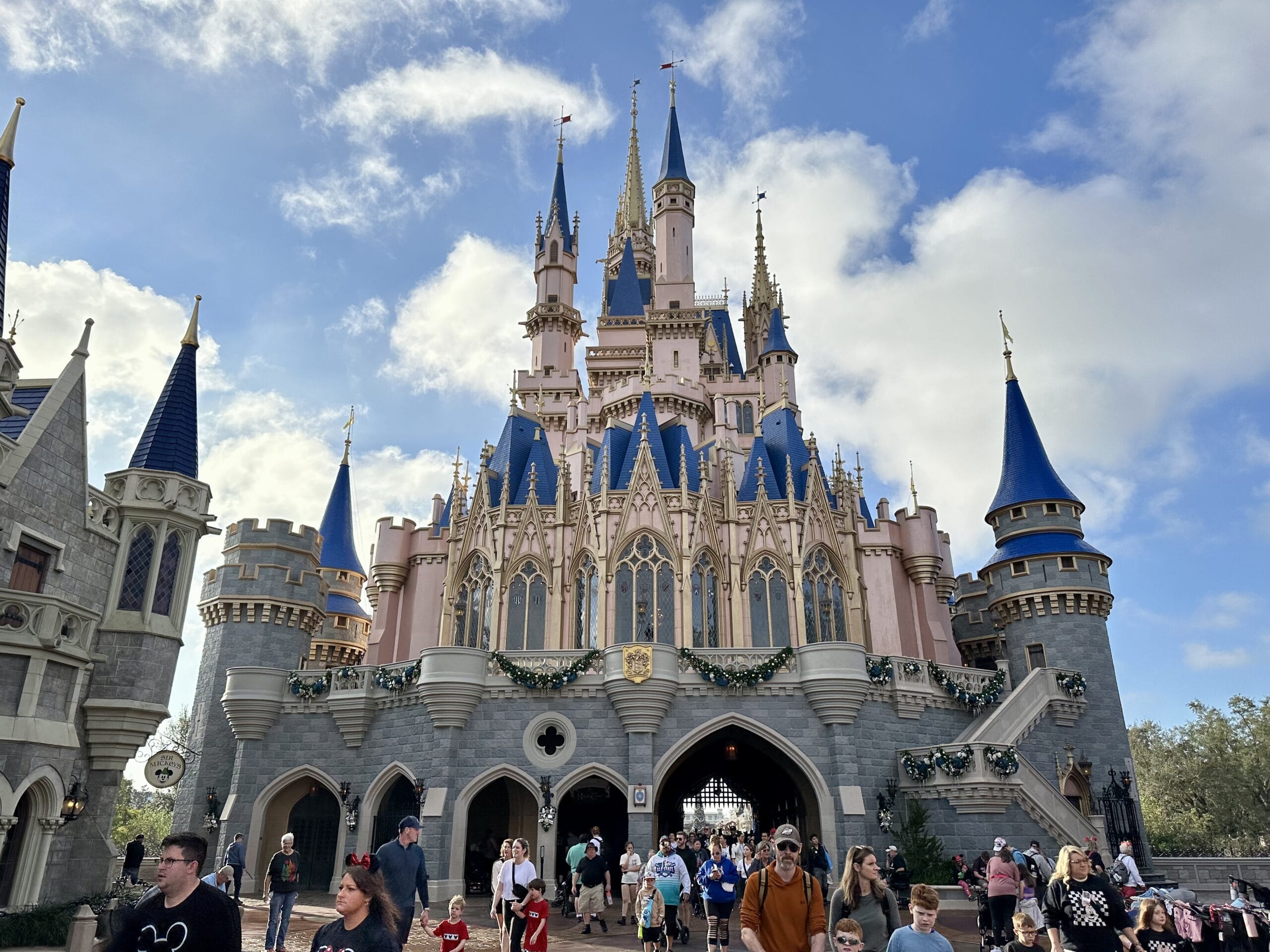 A large castle with blue spires and decorative details under a partly cloudy sky, surrounded by people walking.