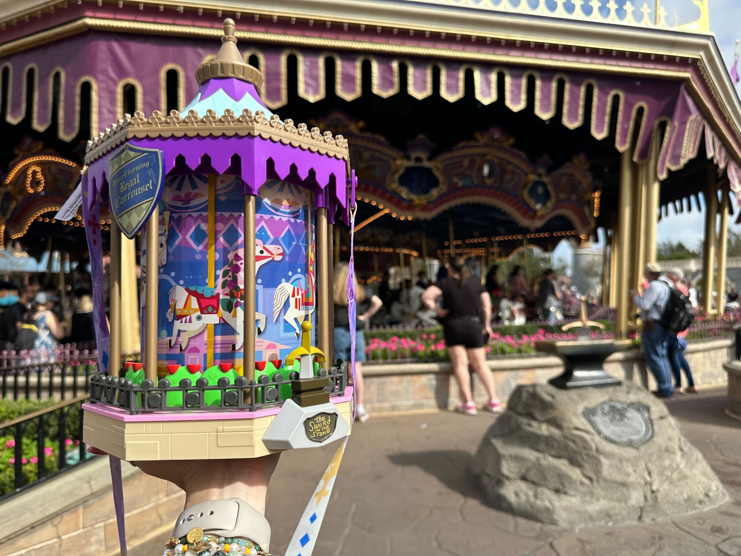 Hand holding a carousel-themed popcorn bucket in front of the Prince Charming Regal Carrousel at Magic Kingdom.