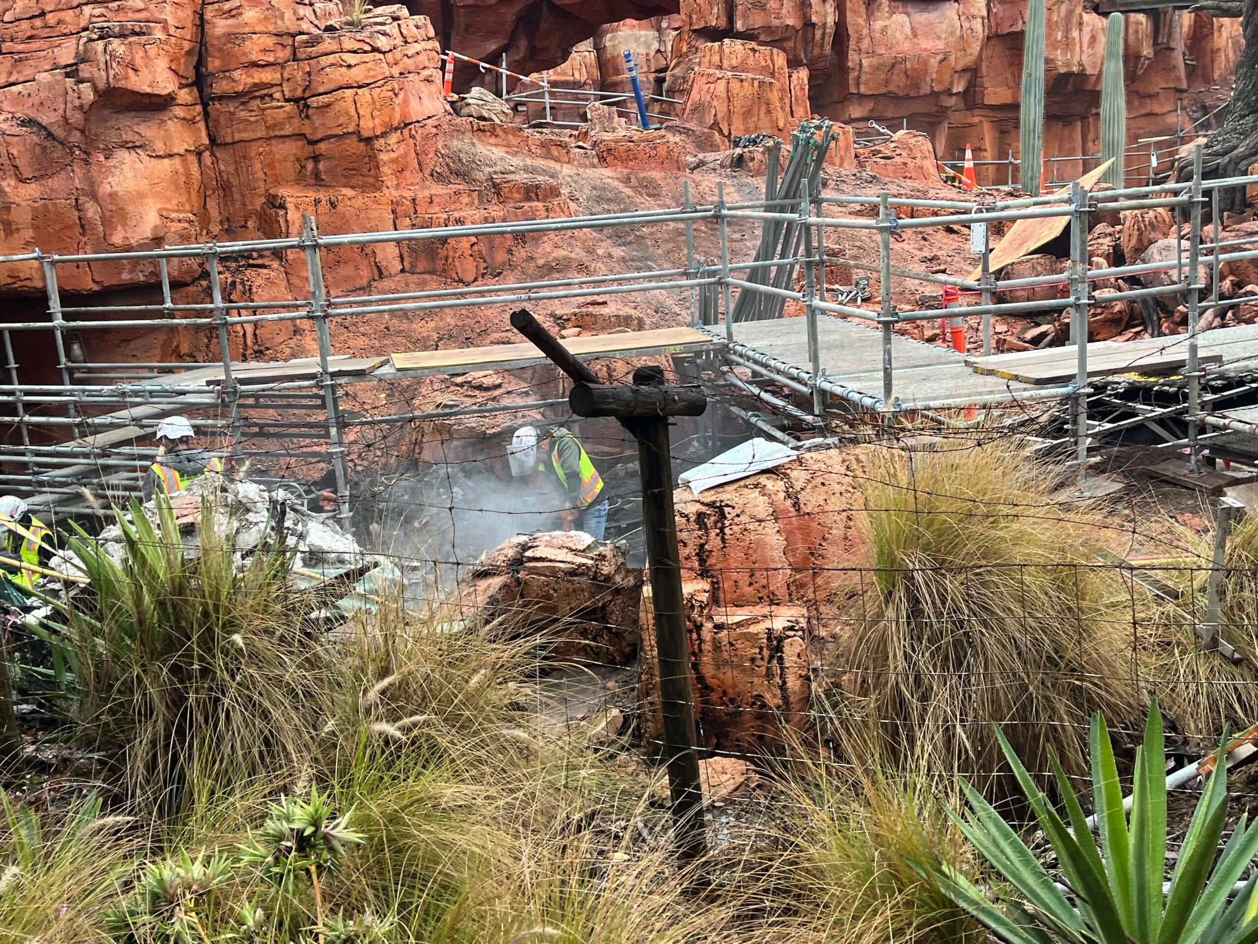 Construction workers at a site with rocky terrain and scaffolding, surrounded by dry vegetation.