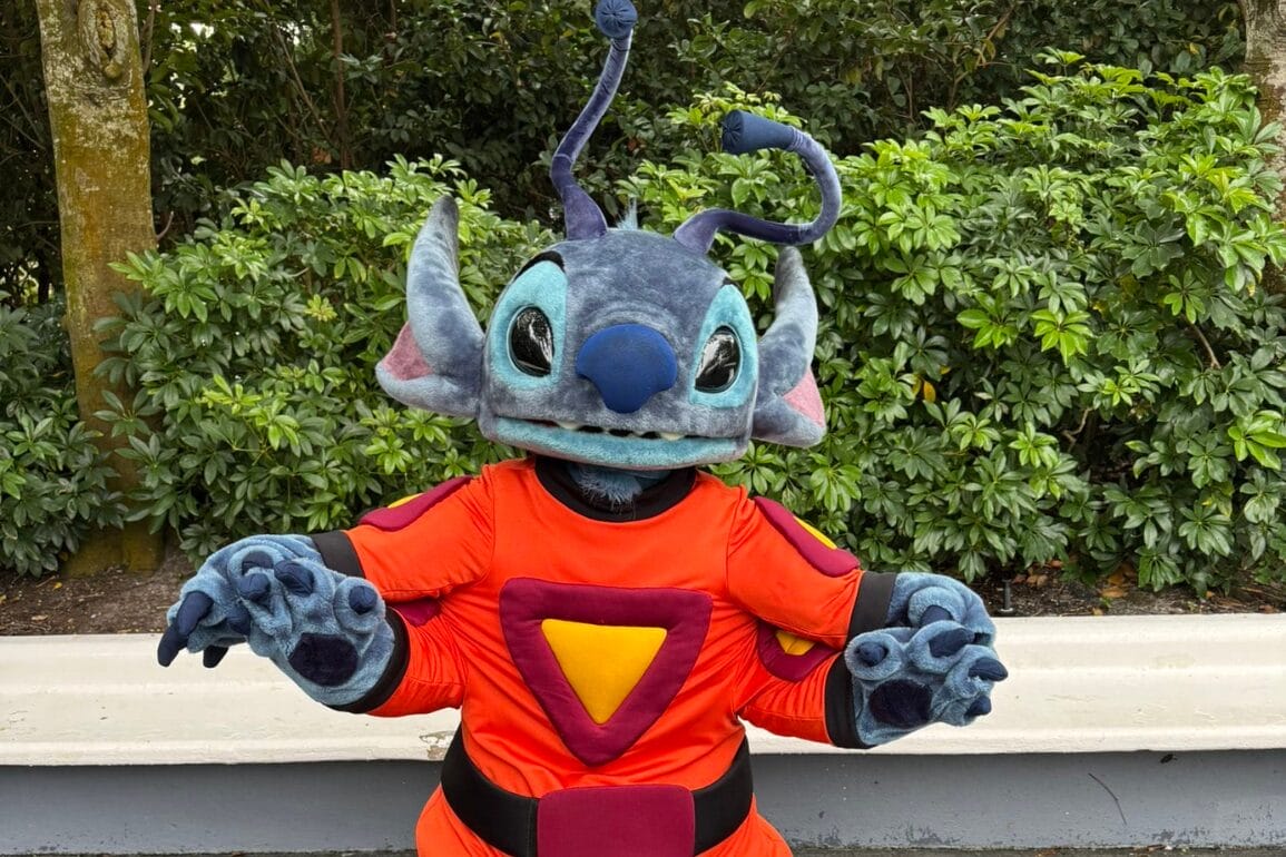 A person in an Alien Stitch costume, donning an orange space suit, poses outdoors amid lush greenery for a magical Meet & Greet experience.