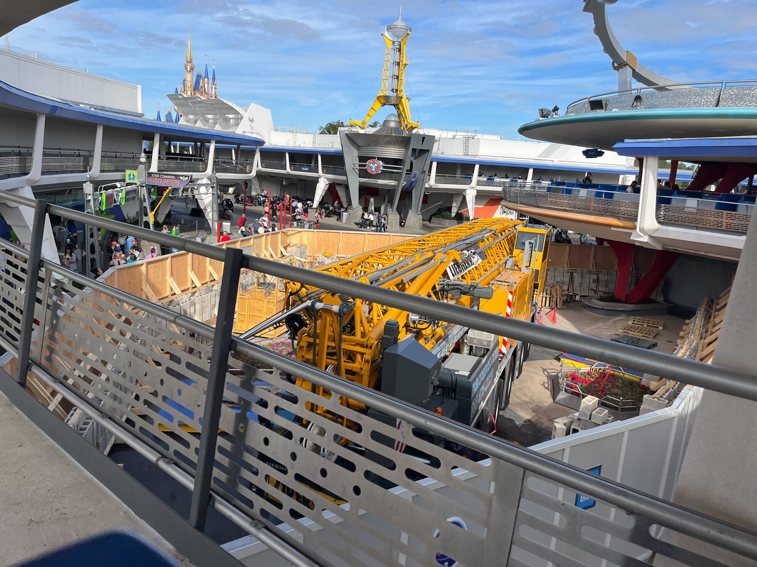 Construction work at Magic Kingdom's open space features a large yellow crane and various materials, with people walking nearby and futuristic Astro Orbiter structures in the background, signaling an exciting refurbishment underway.