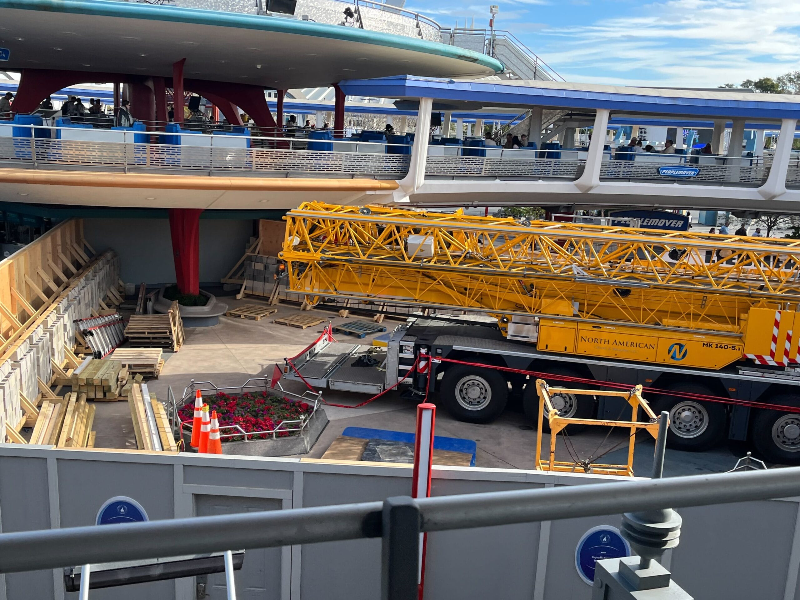 A construction site with a large yellow crane on a truck, surrounded by barriers and building materials, hints at an ongoing refurbishment. In the background, an elevated walkway and people are visible near the Magic Kingdom's Astro Orbiter.