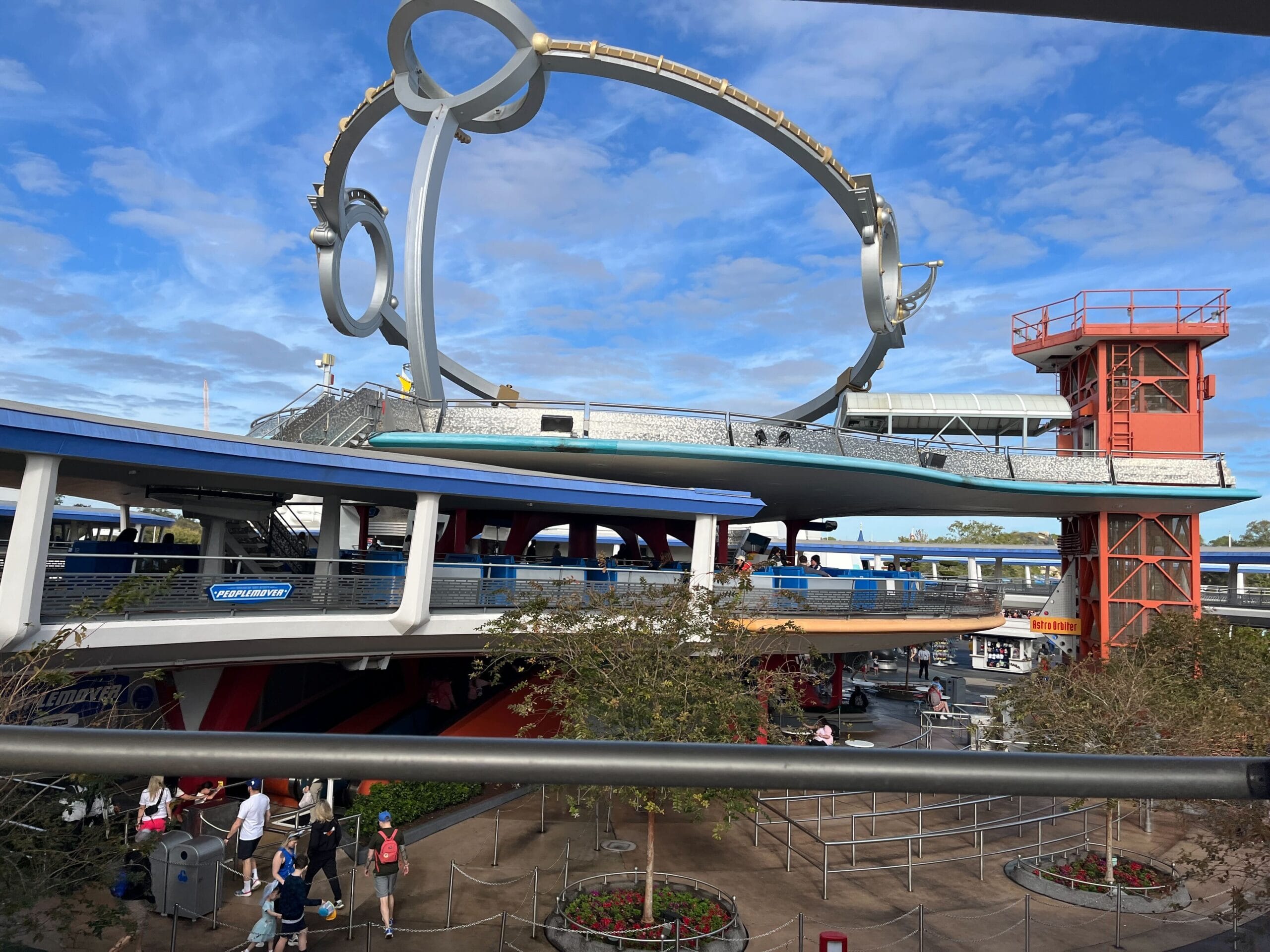 The outdoor futuristic theme park structure features the Astro Orbiter, a large metallic circular ride above. A red and white building stands on the right as visitors stroll below under a partly cloudy sky, evoking the enchanting atmosphere of Magic Kingdom.