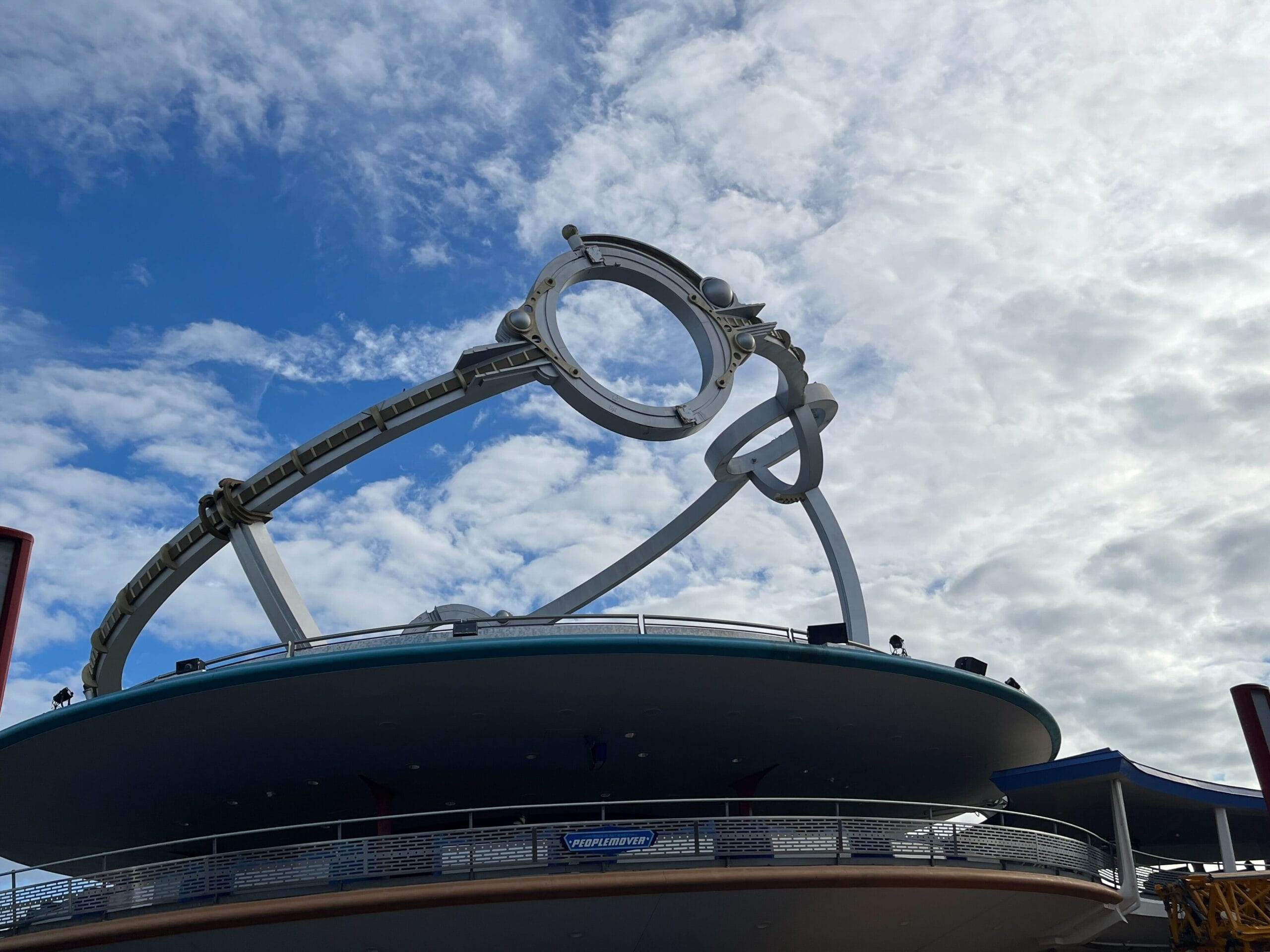 A futuristic, circular metal structure with a sleek design stands against a partly cloudy sky, resembling an Astro Orbiter atop a modern building with a balcony and railing.