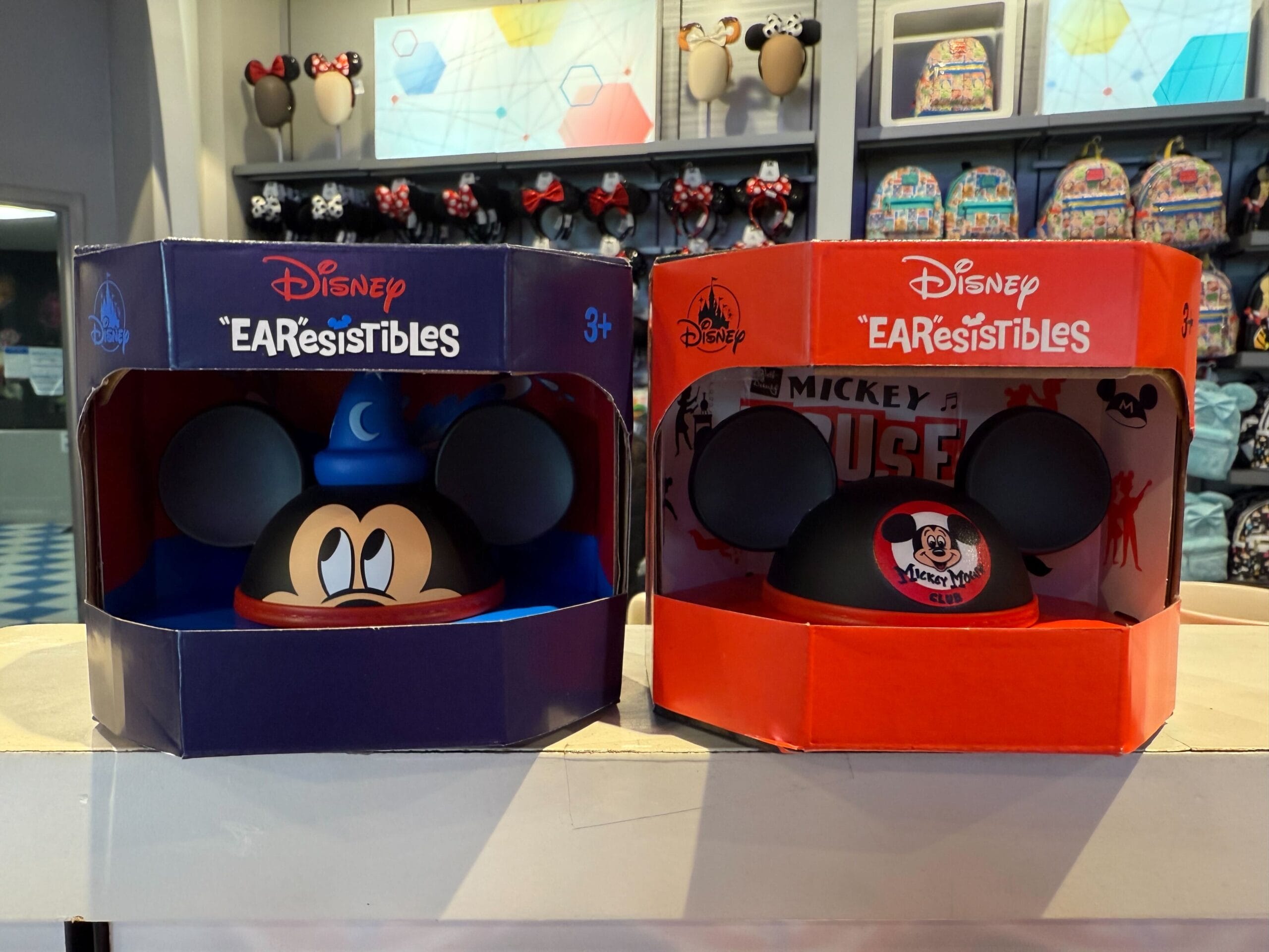 Two boxed Disney "EARSistibles" featuring Mickey Mouse ears are displayed on a store shelf. The left box is blue with a sorcerer design, and the right box is red with a Mickey Mouse Club design.